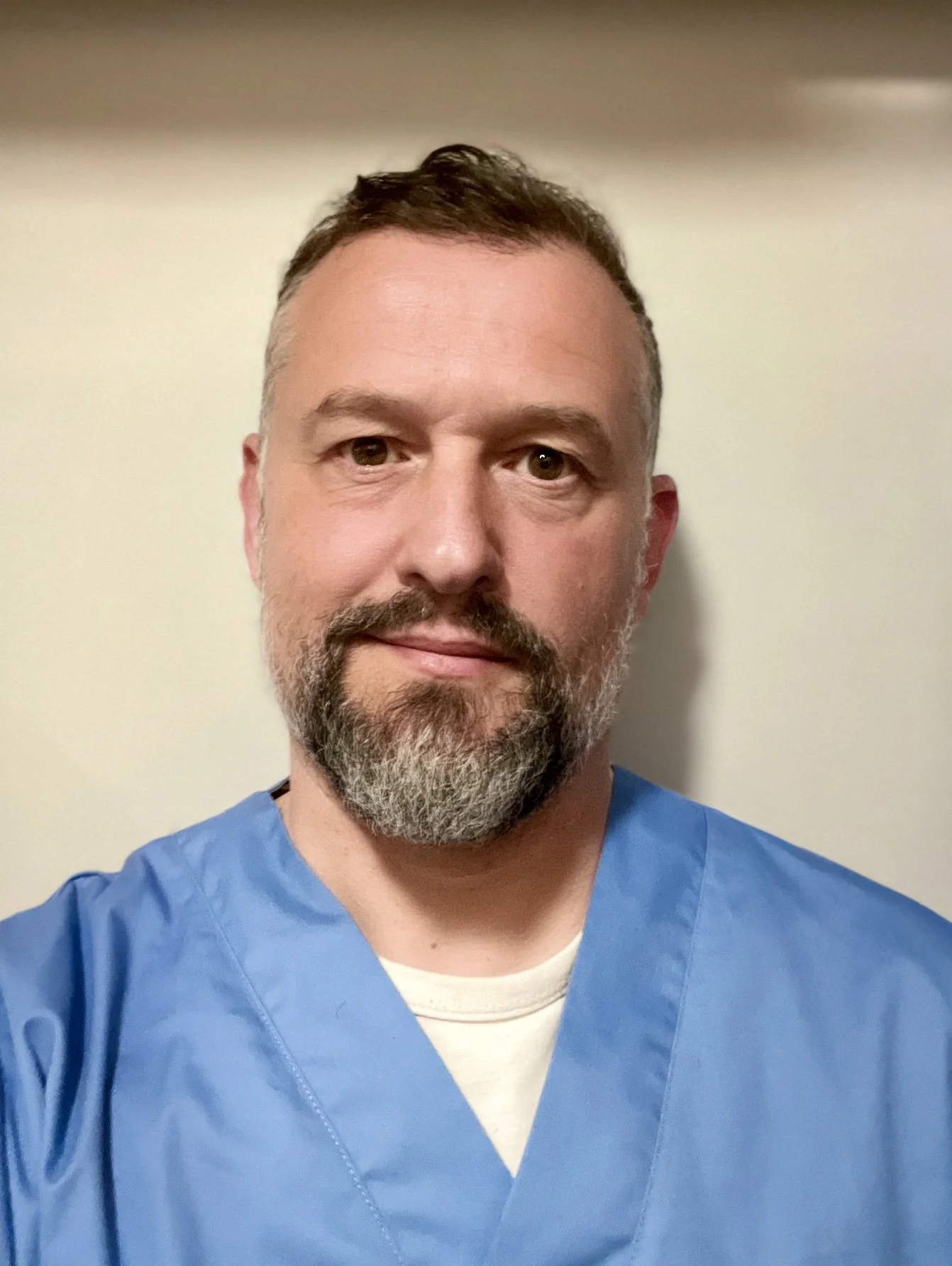 Hombre con barba y cabello castaño, vestido con uniforme médico azul, frente a una pared clara.