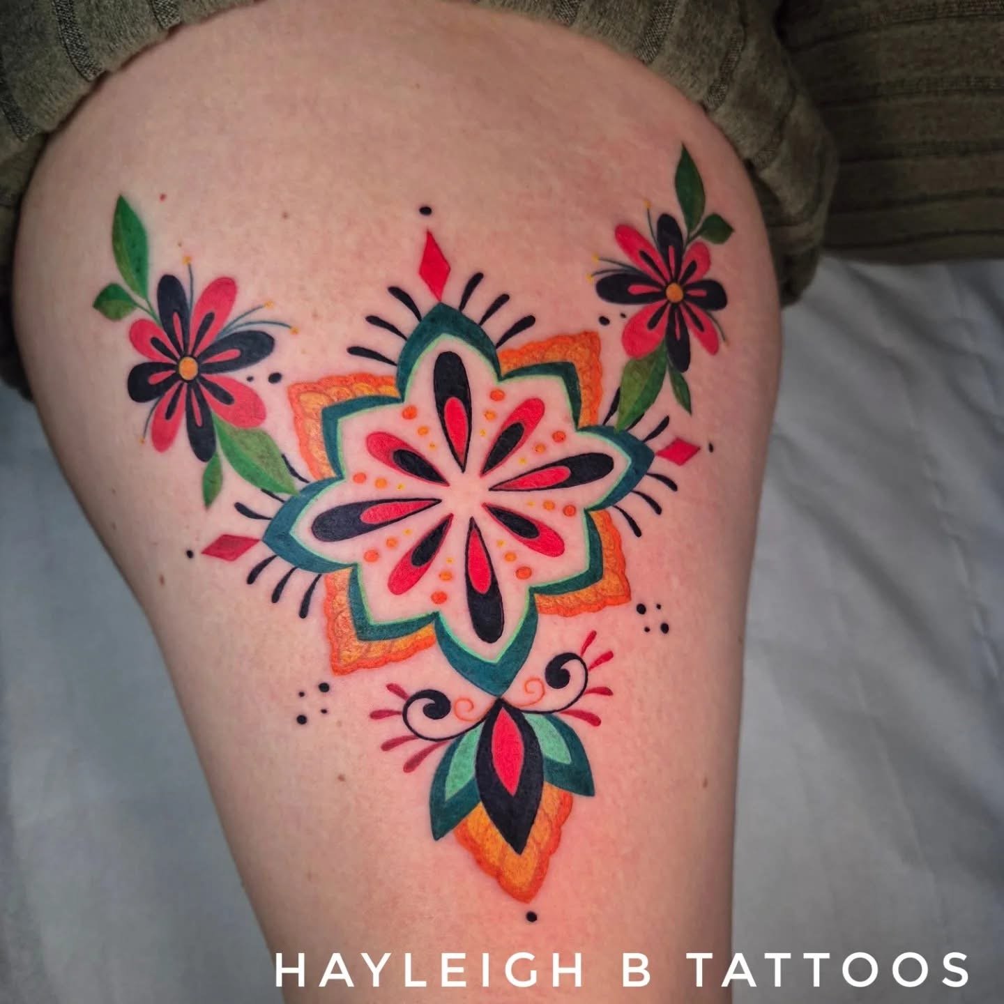 Colorful floral tattoo on the thigh with pink, black, green, orange, and red colors, featuring flowers and decorative patterns. Text overlay reads 'HAYLEIGH B TATTOOS'.