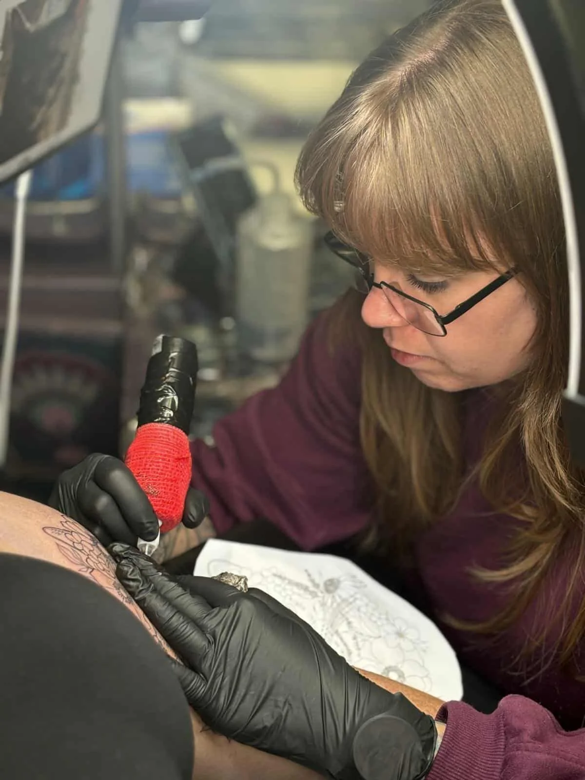 Tattoo artist using a tattoo machine on a client's arm, with the artist wearing black gloves and the client’s arm resting on a surface.