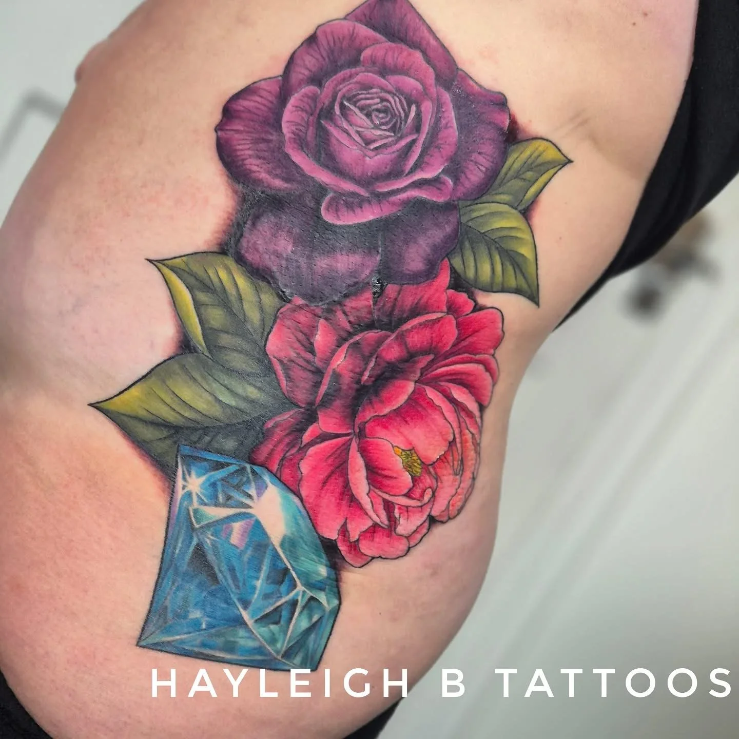 Tattoo of two roses, one purple and one red, with green leaves, and a blue gemstone at the bottom, on a person's arm.