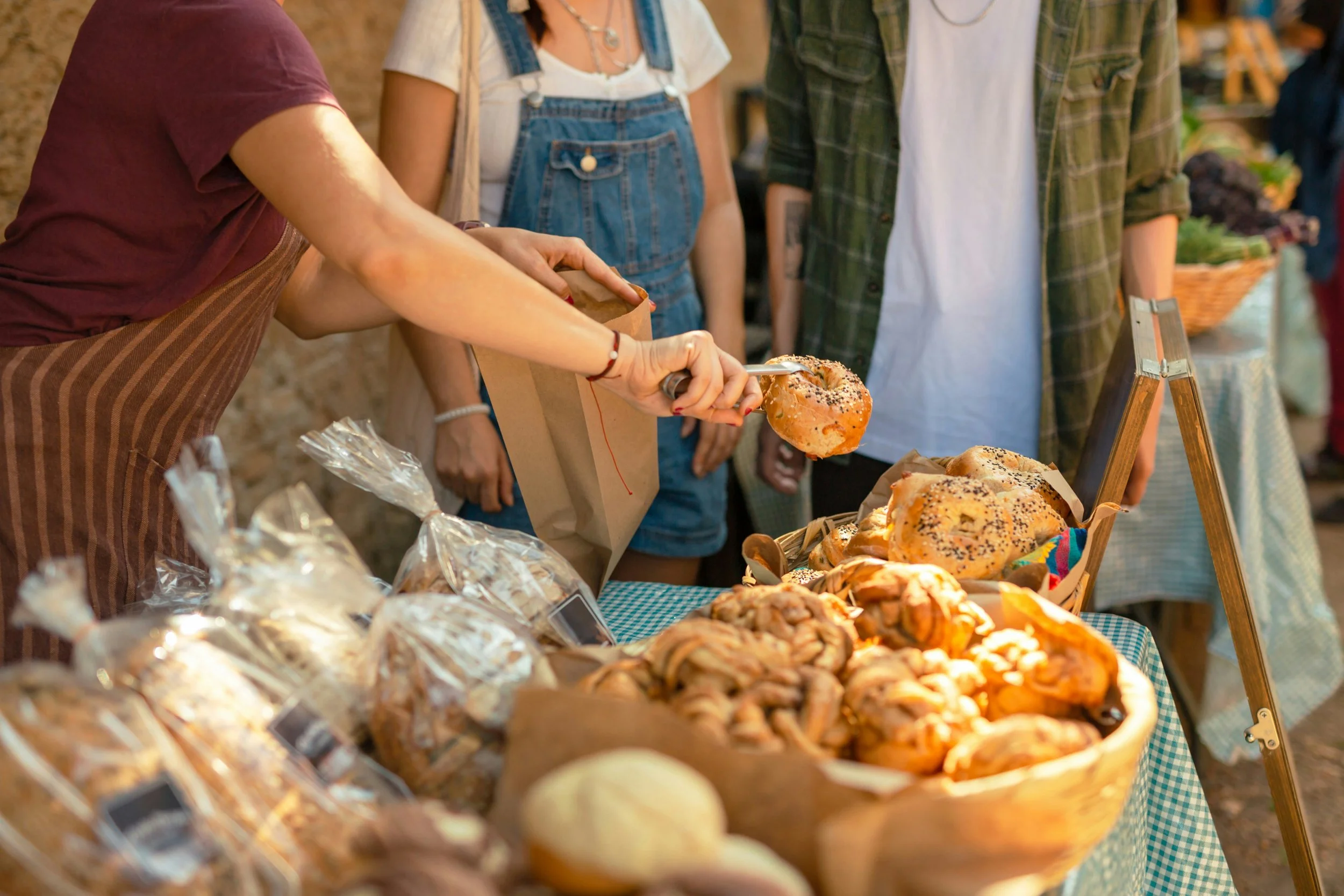 People browsing baked goods at an outdoor market, with pastries and bread on a checkered tablecloth.
