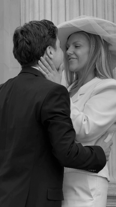 A couple, with the woman dressed as a bride in a white outfit and veil, and the man in a dark suit, share a close, intimate moment, possibly at their wedding, with the woman smiling and touching the man's face.