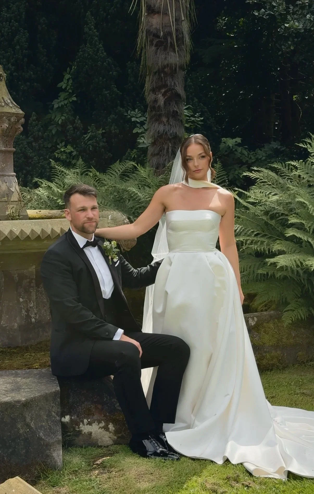 A bride in an elegant white wedding gown with a veil and a groom in a black tuxedo with a bow tie, posing outdoors in a lush green garden with ferns and trees.