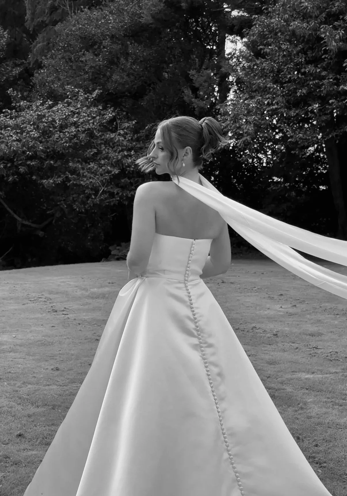 A woman in a strapless wedding gown with a long veil stands outdoors on a grassy area surrounded by trees, facing away from the camera.