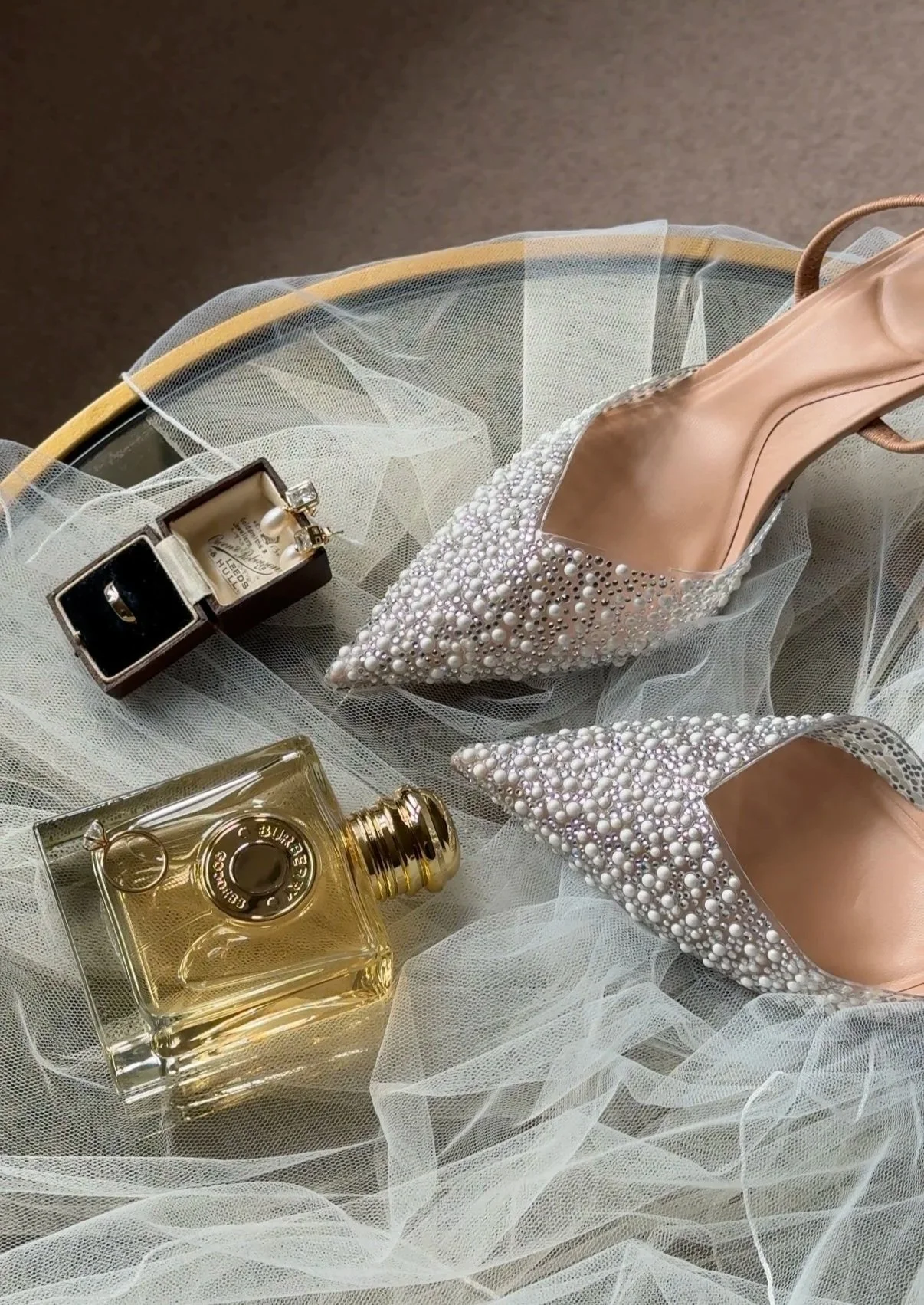 A pair of embellished high-heeled shoes with pearl details, a bottle of perfume, a jewelry box with rings, and a sheer fabric on a surface.