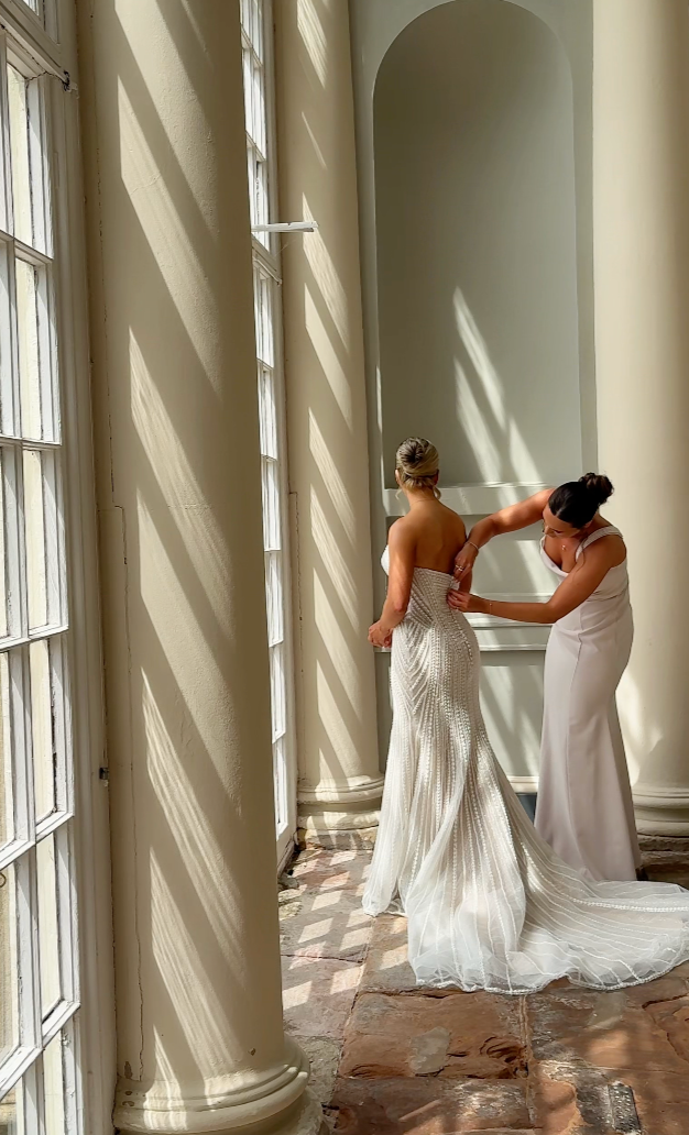 A woman in a strapless wedding dress standing inside near tall windows with sunlight streaming in, as another woman helps her get ready.