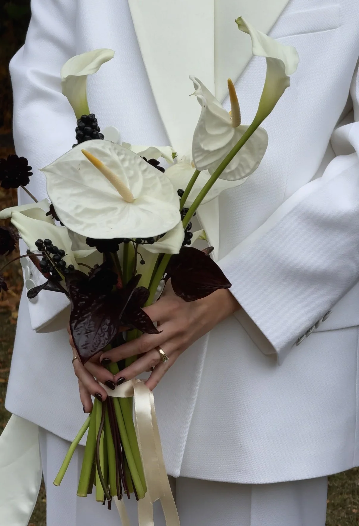A person in white attire holding a bouquet of white calla lilies, dark purple leaves, black berries, and a beige ribbon.