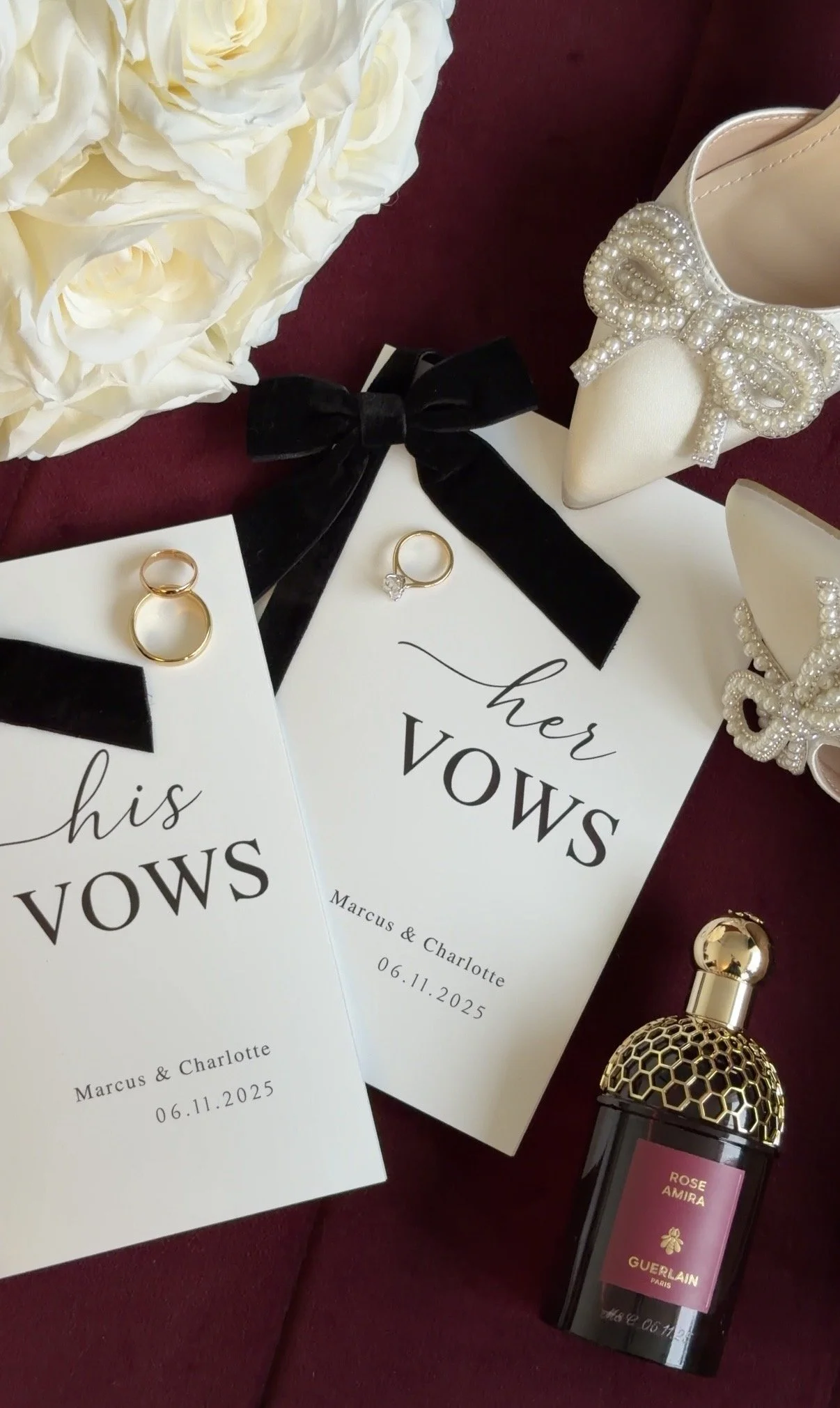 Wedding vow booklets from Marcus & Charlotte with date 06.11.2025, rings, bridal shoes with pearl adornment, perfume bottle, flowers, and accessories on a dark red background.