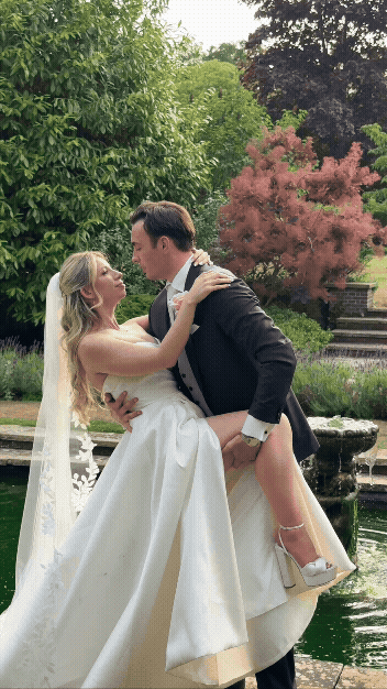 Bride and groom dancing outdoors near a pond with trees in the background, the groom lifting the bride as she wears a white wedding gown and veil