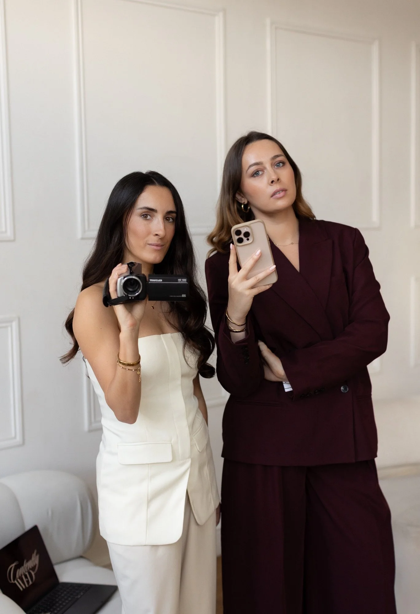 Two women standing indoors, one holding a camera and the other holding a smartphone, both looking at the camera with serious expressions.