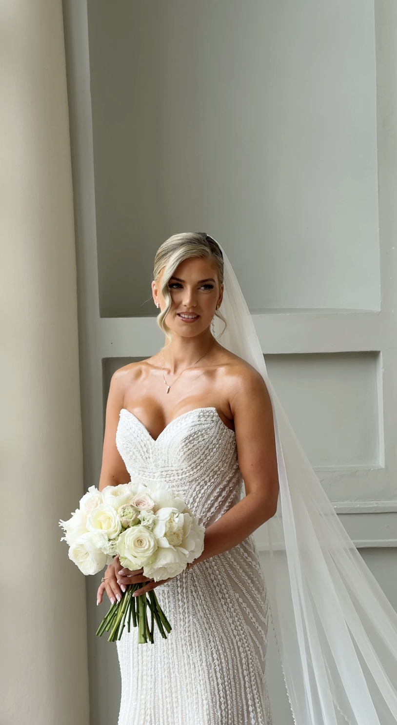 Brunette bride in strapless white wedding dress holding a bouquet of white roses and peonies, standing near a window with natural light, wearing a veil and necklace.