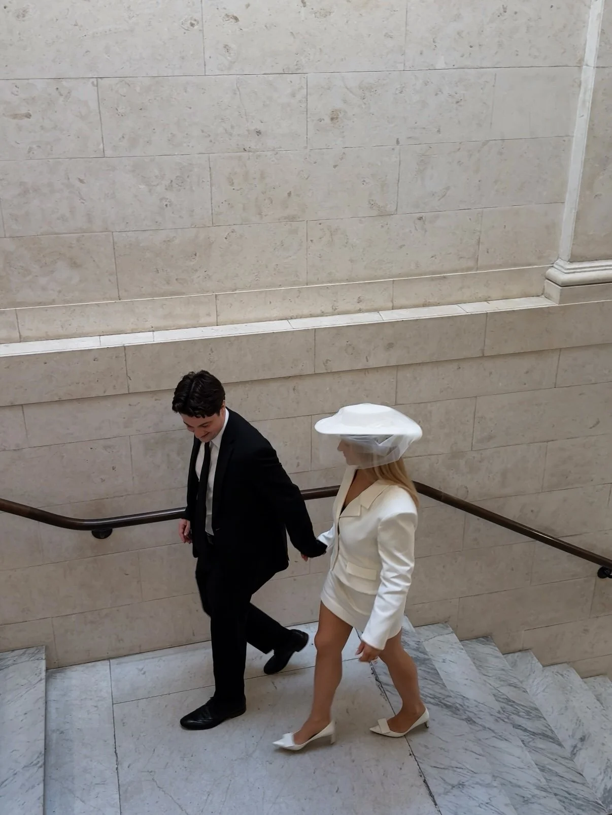 A man in a black tuxedo holding hands with a woman in a white blazer dress, thigh-high white heels, and a wide-brimmed white hat, walking up a marble staircase in an elegant building.