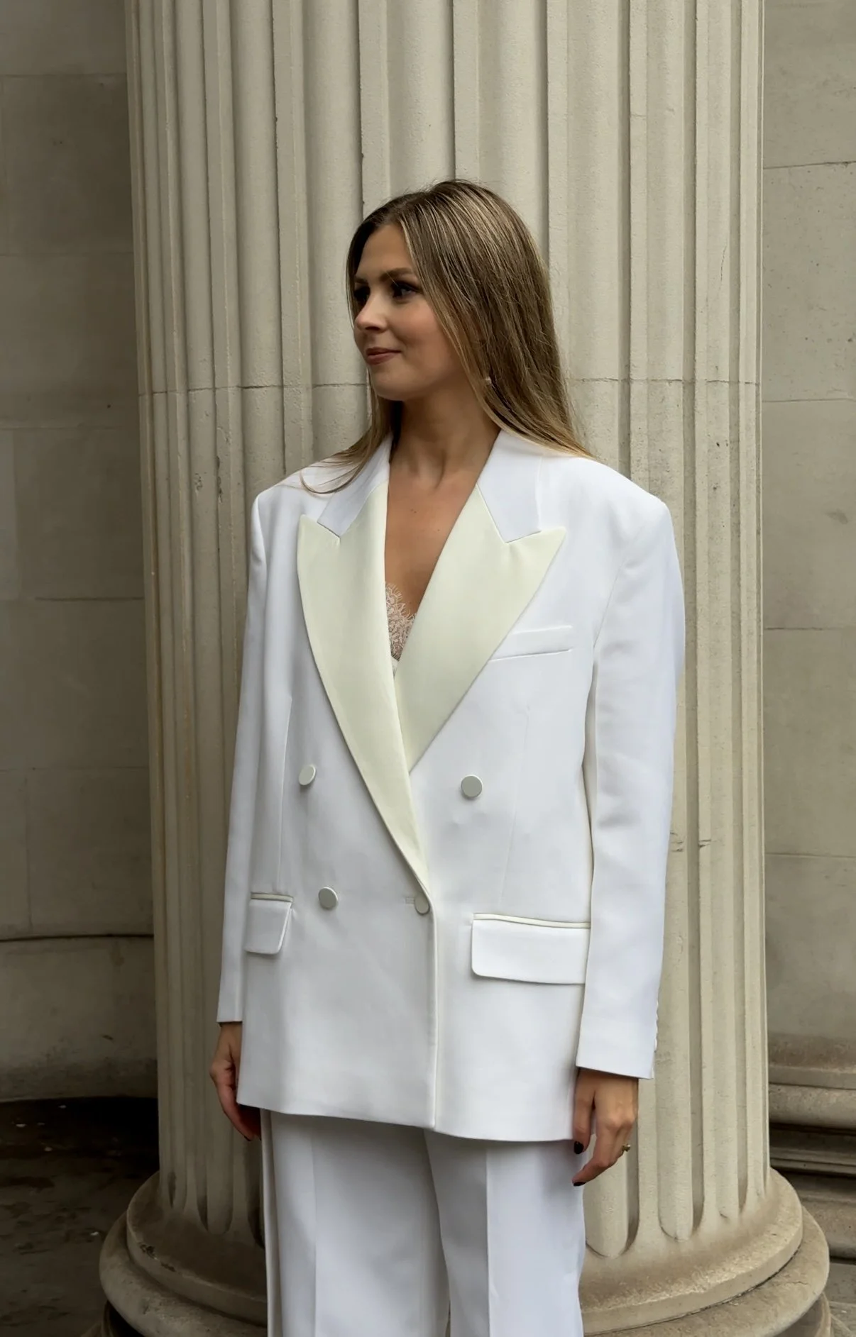 Woman in white pantsuit with beige lapel standing next to a classical column.