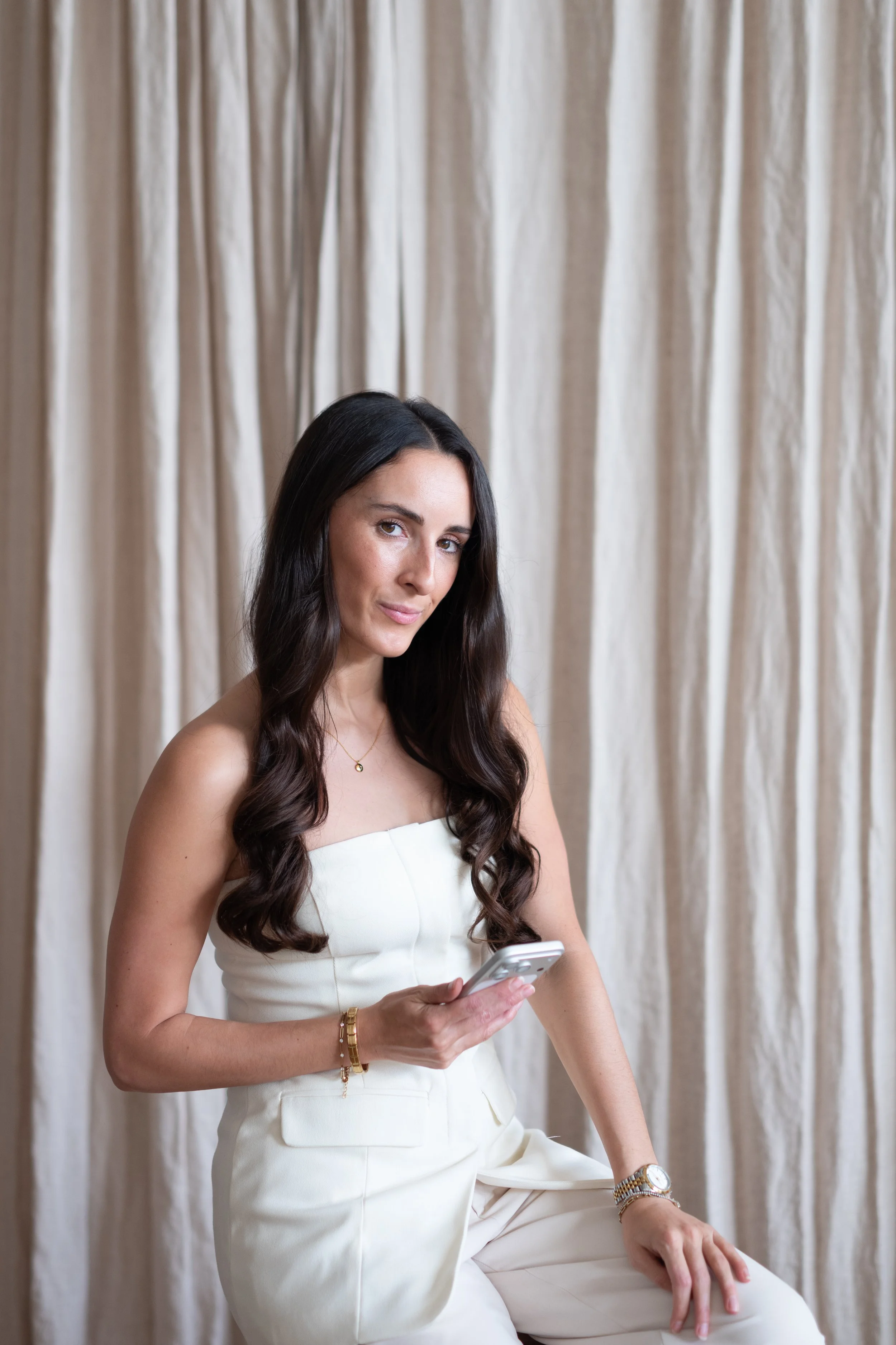 A woman with long dark wavy hair wearing a white strapless top and matching pants, holding a smartphone, sitting against a cream-colored curtain background.