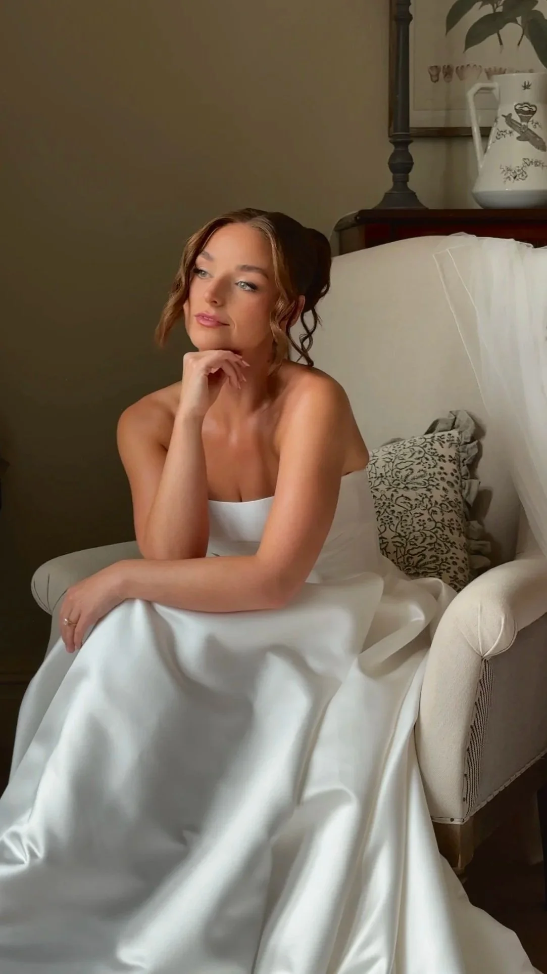 A woman with wavy brown hair and fair skin sits on a cream-colored armchair, wearing a white satin dress, resting her chin on her hand and looking thoughtfully to the side. The background includes a dark wood piece of furniture, a large framed artwork, and a white pitcher on top.