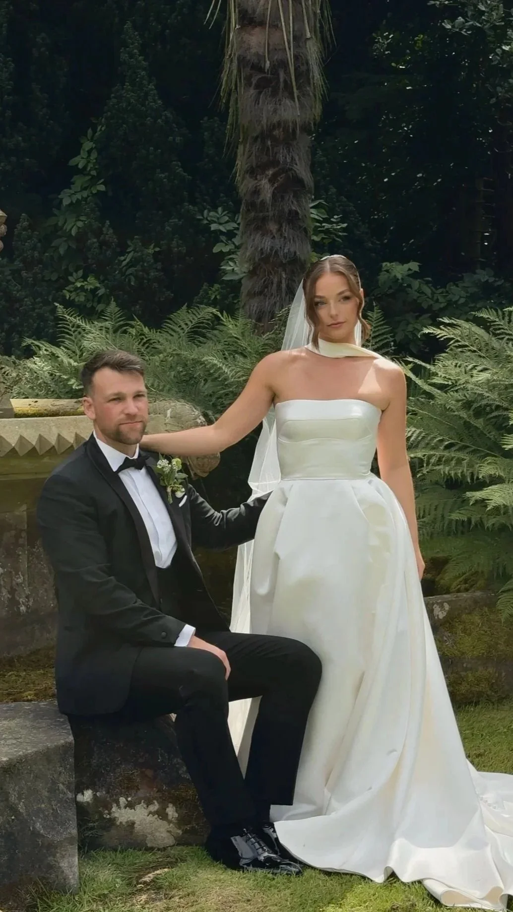 A bride and groom in wedding attire posing outdoors surrounded by greenery and a palm tree.