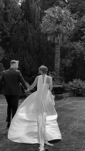 A bride and groom walking hand in hand outdoors on a wedding day, surrounded by trees and greenery.