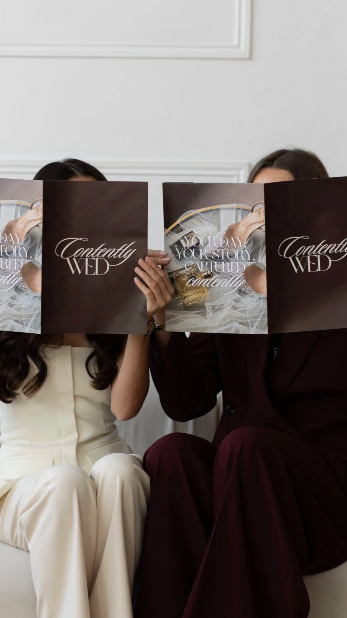 Two women are sitting on a white couch holding up a wedding magazine, which covers their faces. One woman is wearing cream-colored pants and a matching blazer, and the other is in a dark burgundy suit. The magazine has text that reads 'Contentedly Wed' and 'Your Day, Your Story, Captured Contentedly.' The background features a white wall with decorative molding.