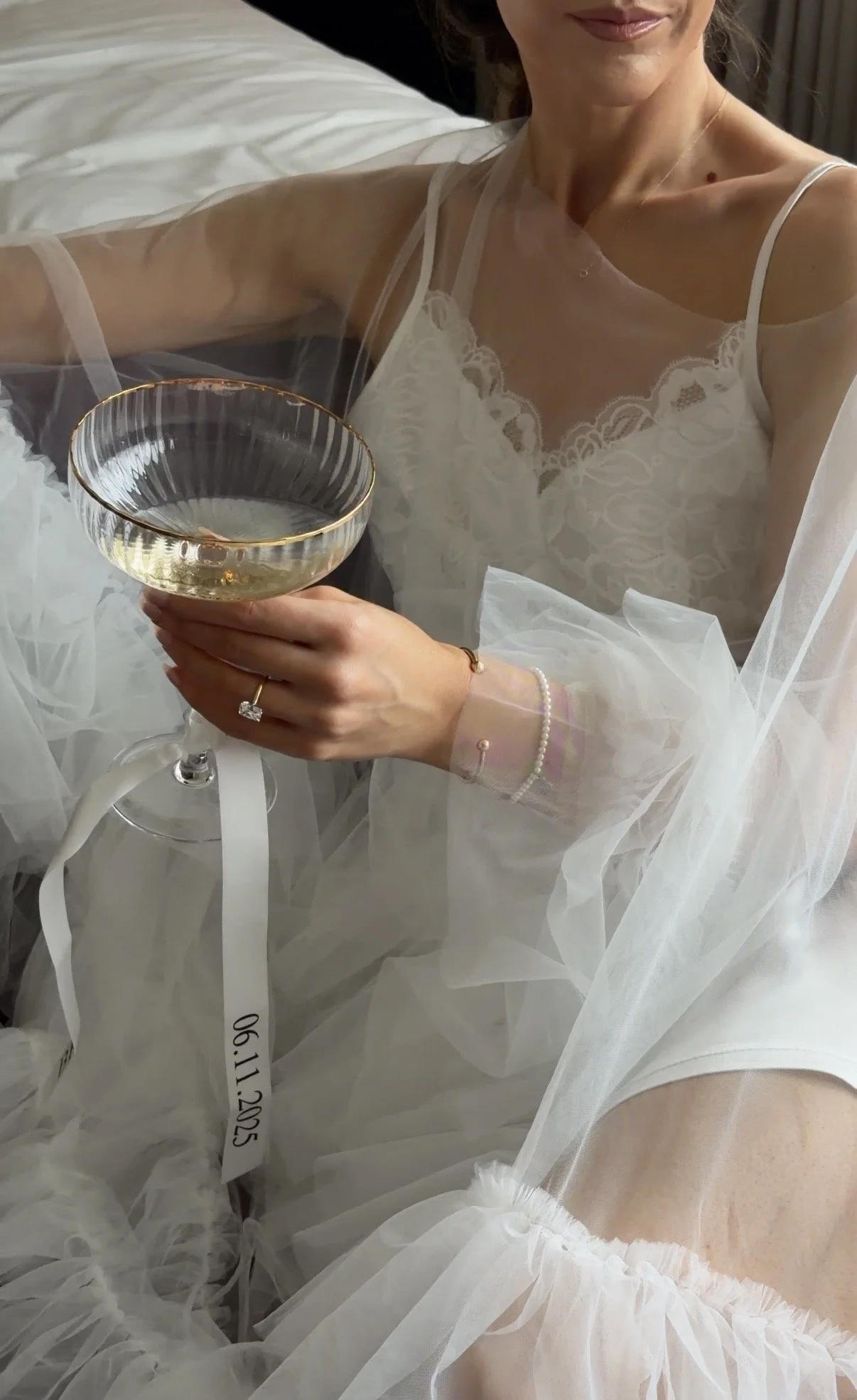 Woman in white lace dress and sheer robes holding a champagne coupe glass, sitting on a bed.