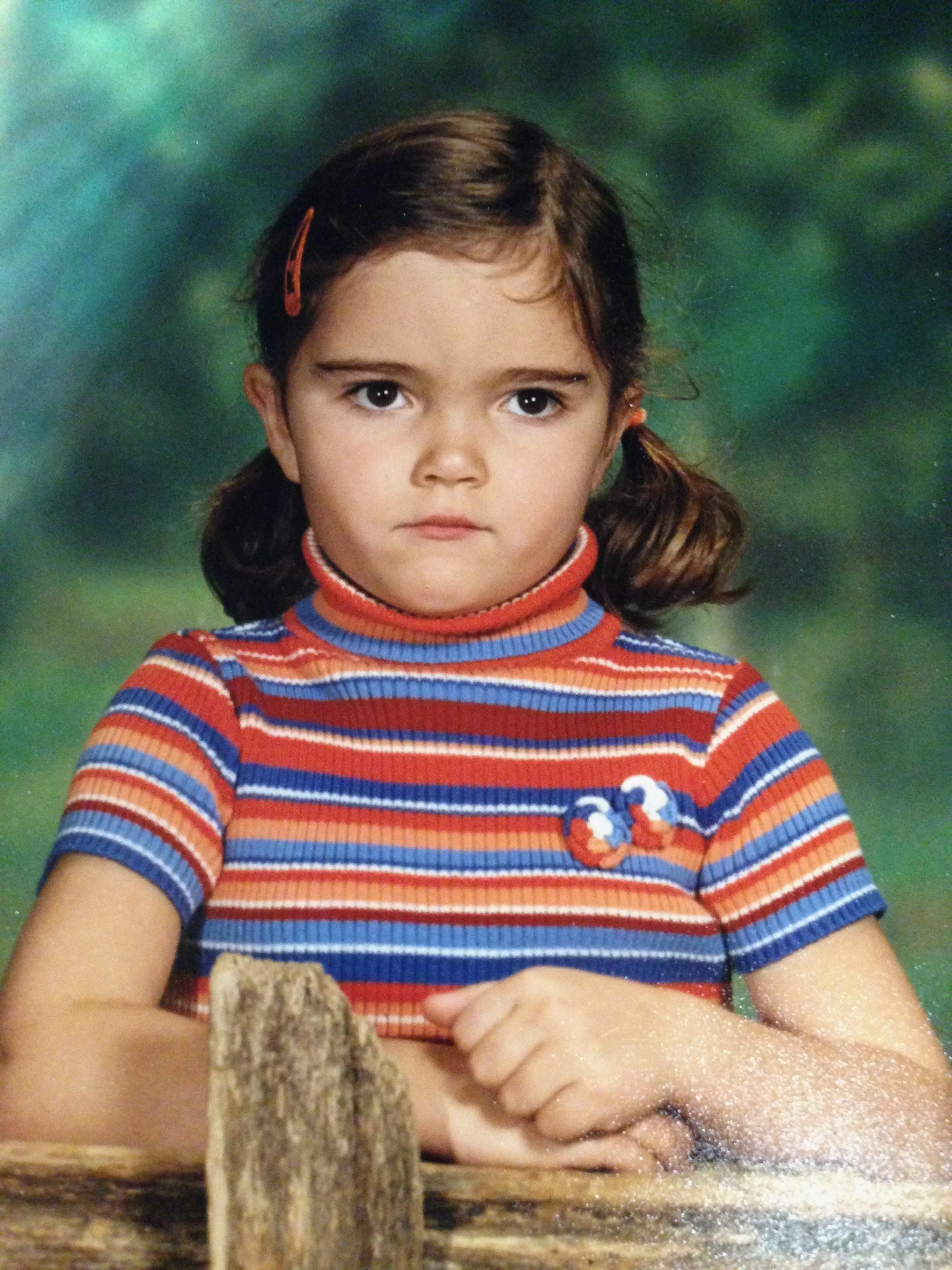 A young girl with dark hair in pigtails wearing a colorful striped turtleneck sweater, sitting outdoors with a green blurred background.