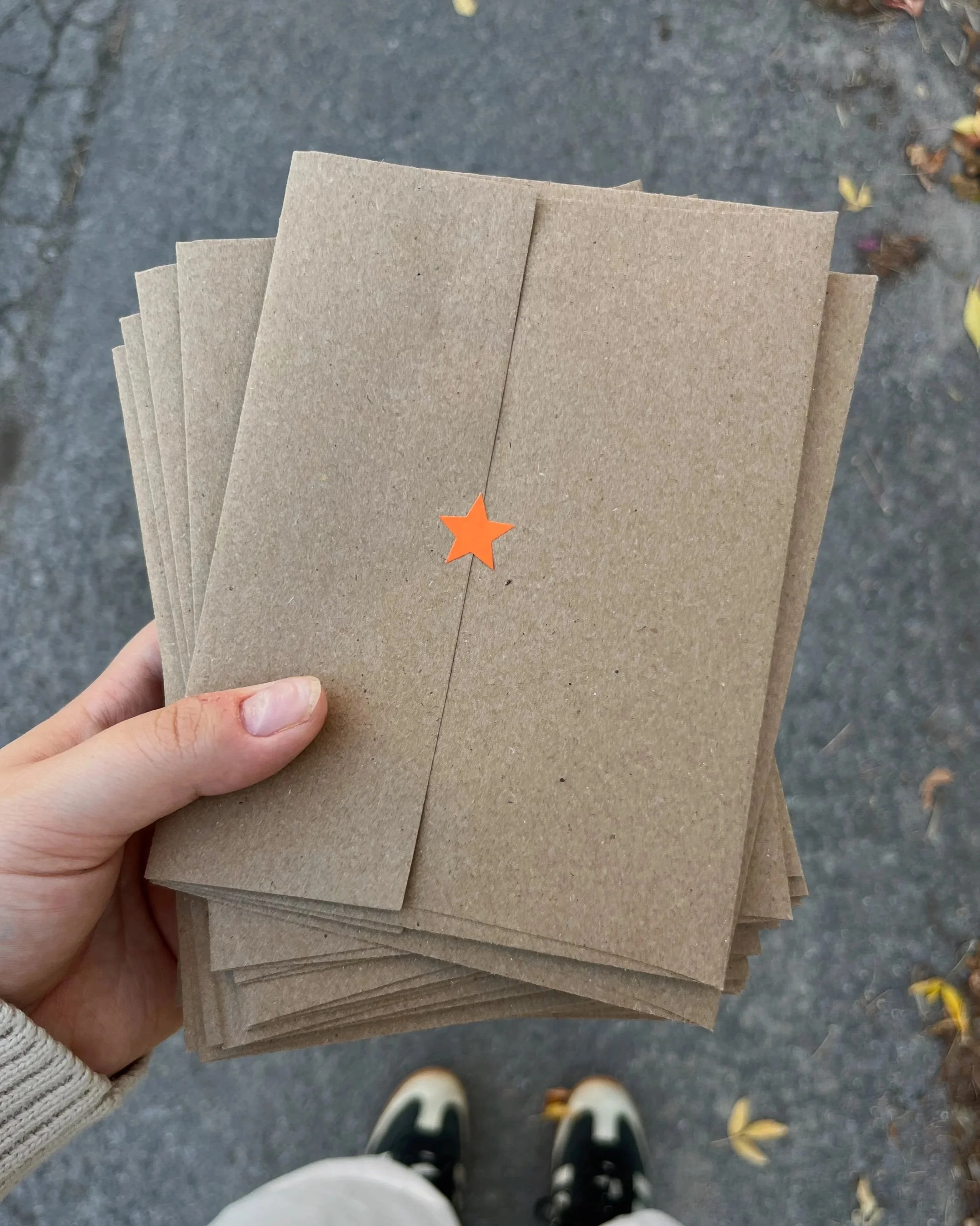 A hand holding a bunch of brown paper napkins or envelopes with an orange star sticker in the center, viewed from above while standing on a sidewalk with fallen yellow leaves.