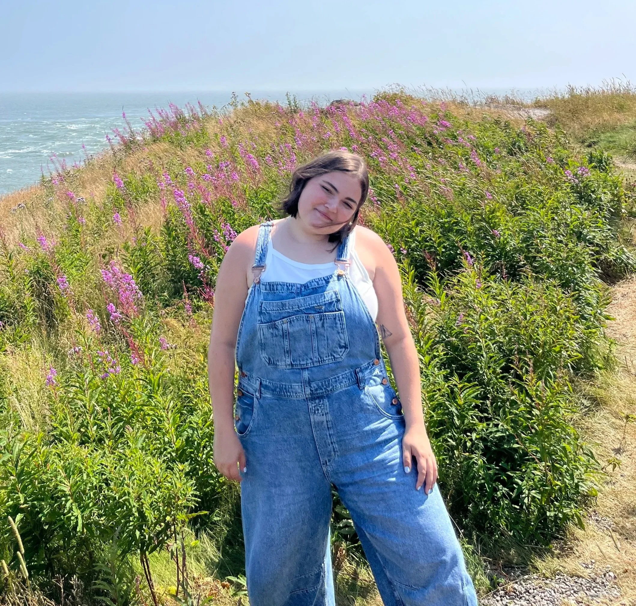 A young woman with dark hair, wearing a white top and denim overalls, stands on a path beside colorful purple flowers with the ocean and sky in the background.