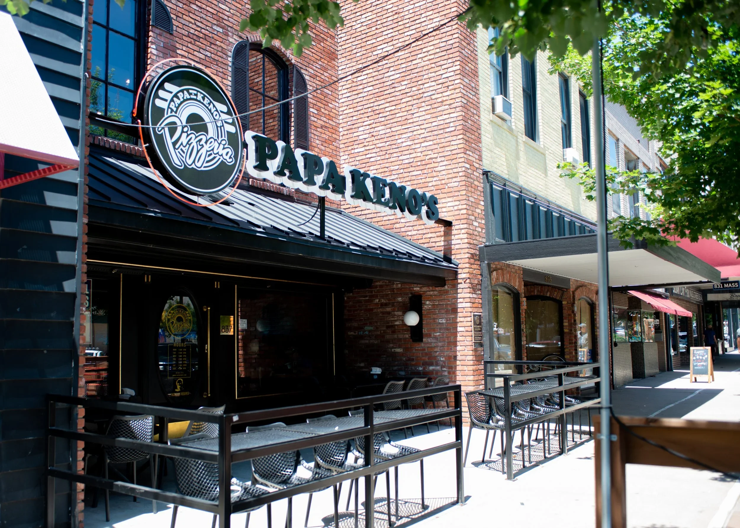 Street view of Papá Kenos pizzeria on a sunny day, with outdoor seating, brick building, and green trees lining the sidewalk.