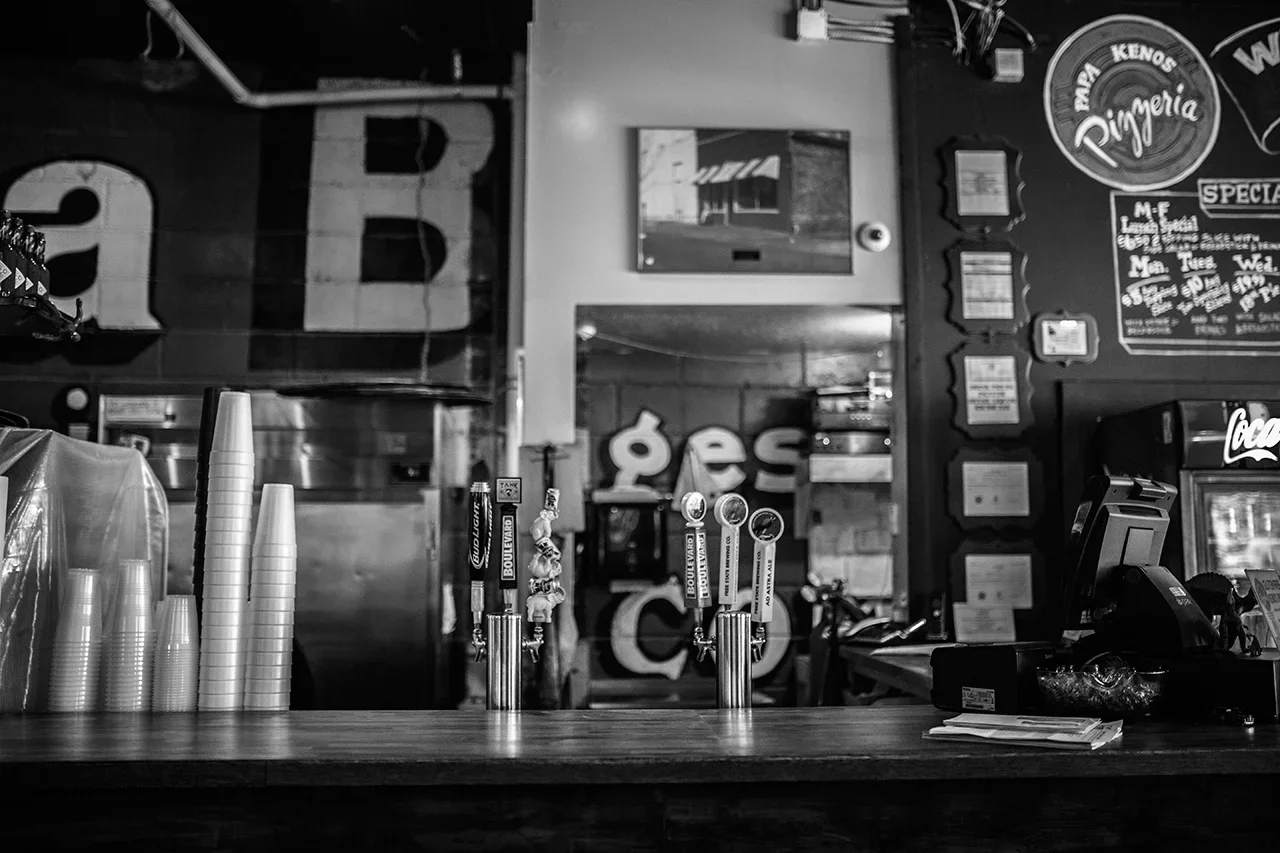 Inside a bar or restaurant with beer tap handles, stacked plastic cups, a cash register, a television mounted on the wall, and various signs and menu boards in the background.