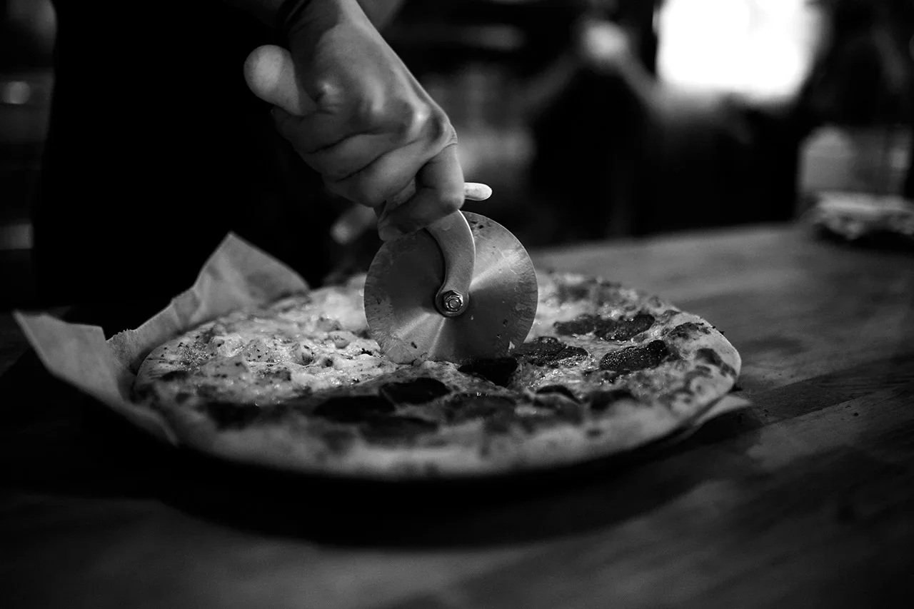 A person cutting a pizza with a pizza cutter on a wooden table in black and white.