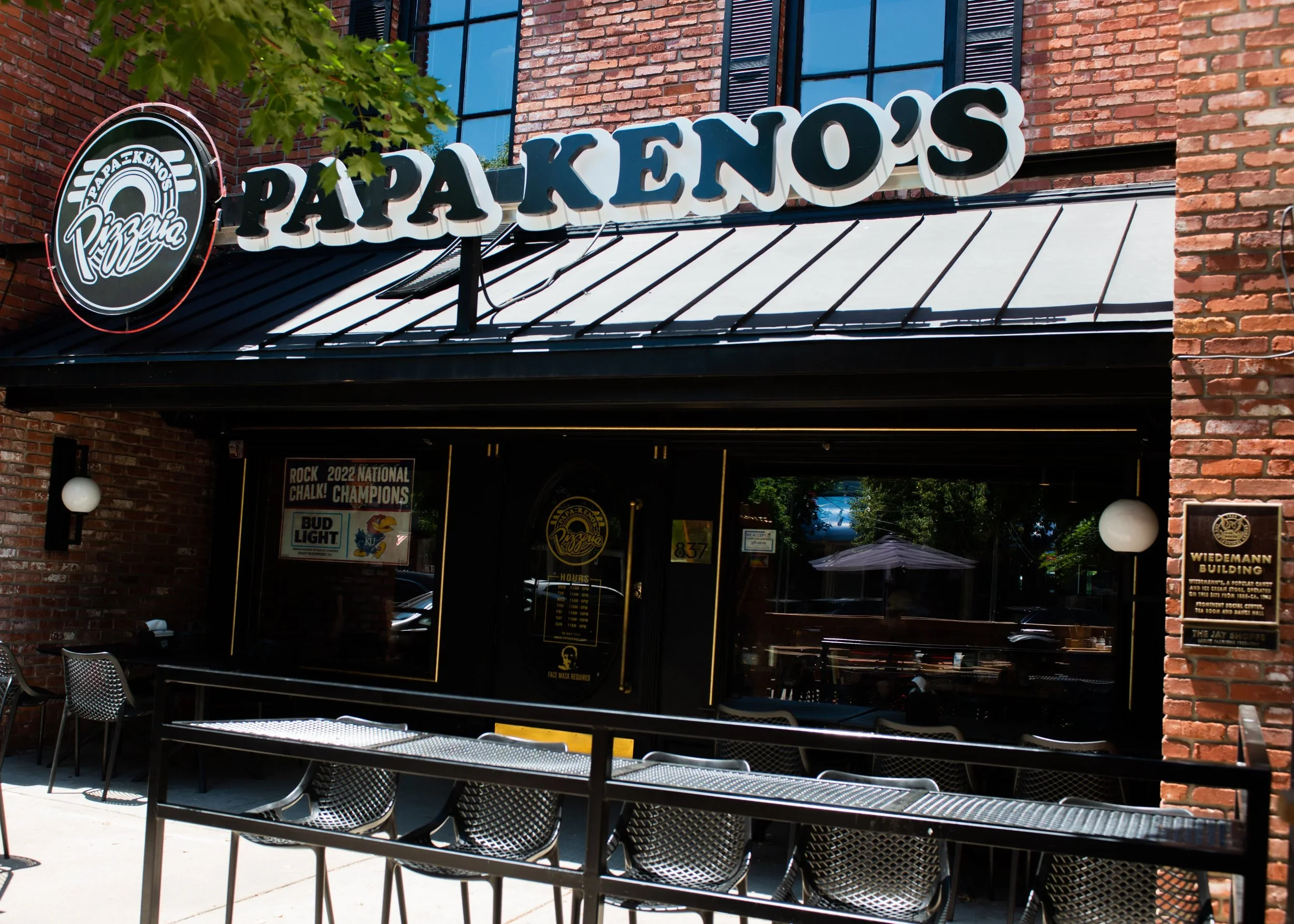 Exterior of Papa Kenos restaurant with a black awning, large white sign, and outdoor seating area with metal chairs and tables in front of a brick building.