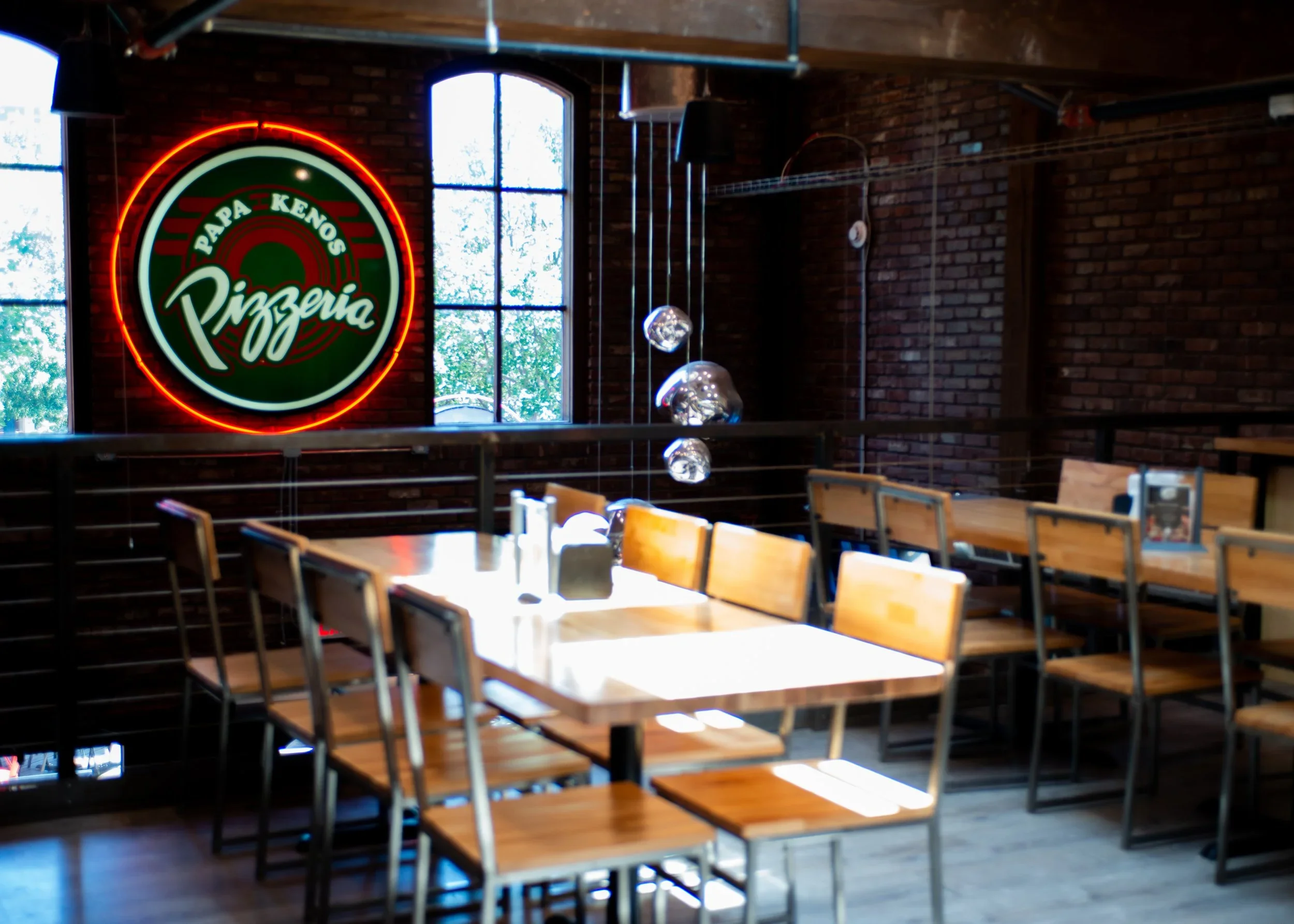 Interior of a pizza restaurant with wooden tables and chairs, brick walls, large windows, a neon sign that reads 'Papa Kenos Pizzeria', and modern hanging light fixtures.