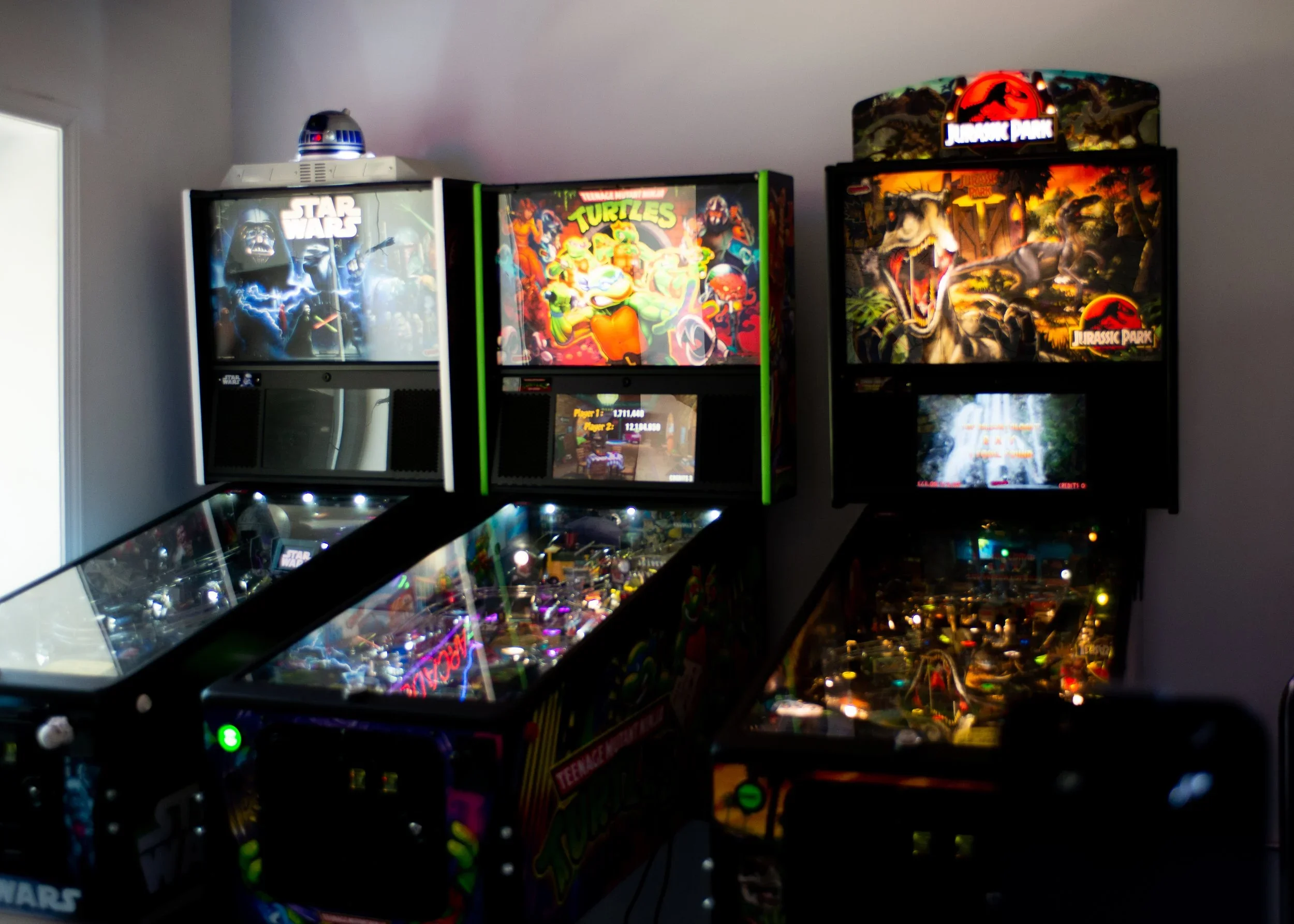 Three pinball machines with colorful artwork, including Jurassic Park and Teenage Mutant Ninja Turtles, in a dimly lit room with a window on the left.