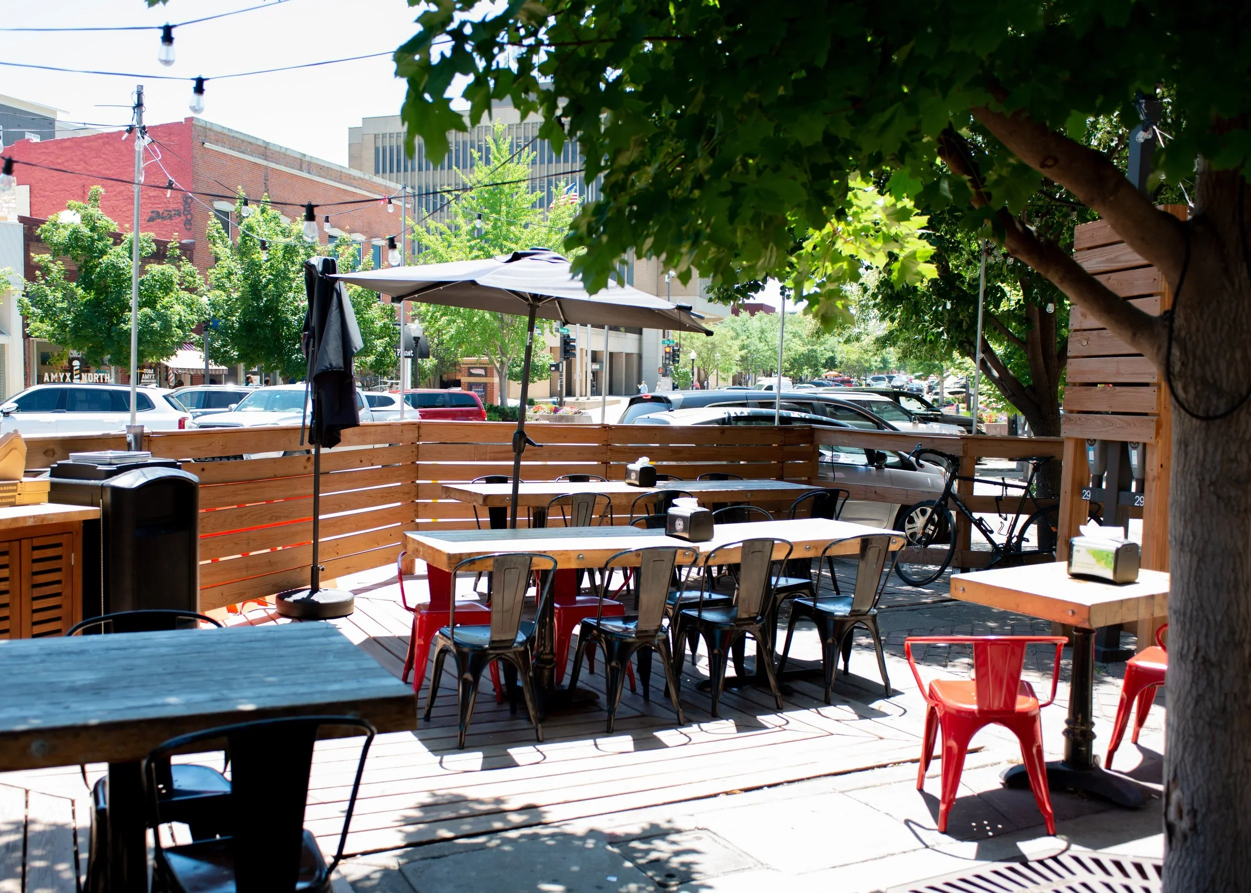 Outdoor patio with tables, chairs, umbrellas, a tree, and string lights, in a city setting with parked cars and buildings in the background.