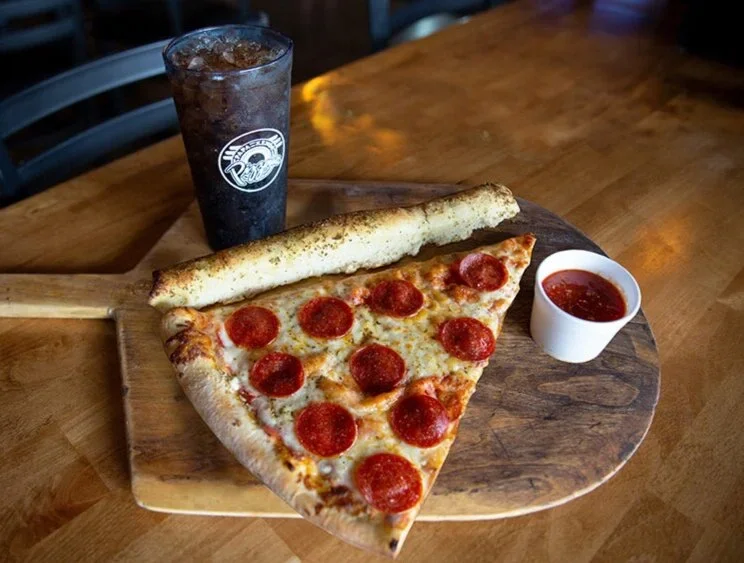 A slice of pepperoni pizza with a breadstick, a cup of marinara sauce, and a glass of soda on a wooden serving board.