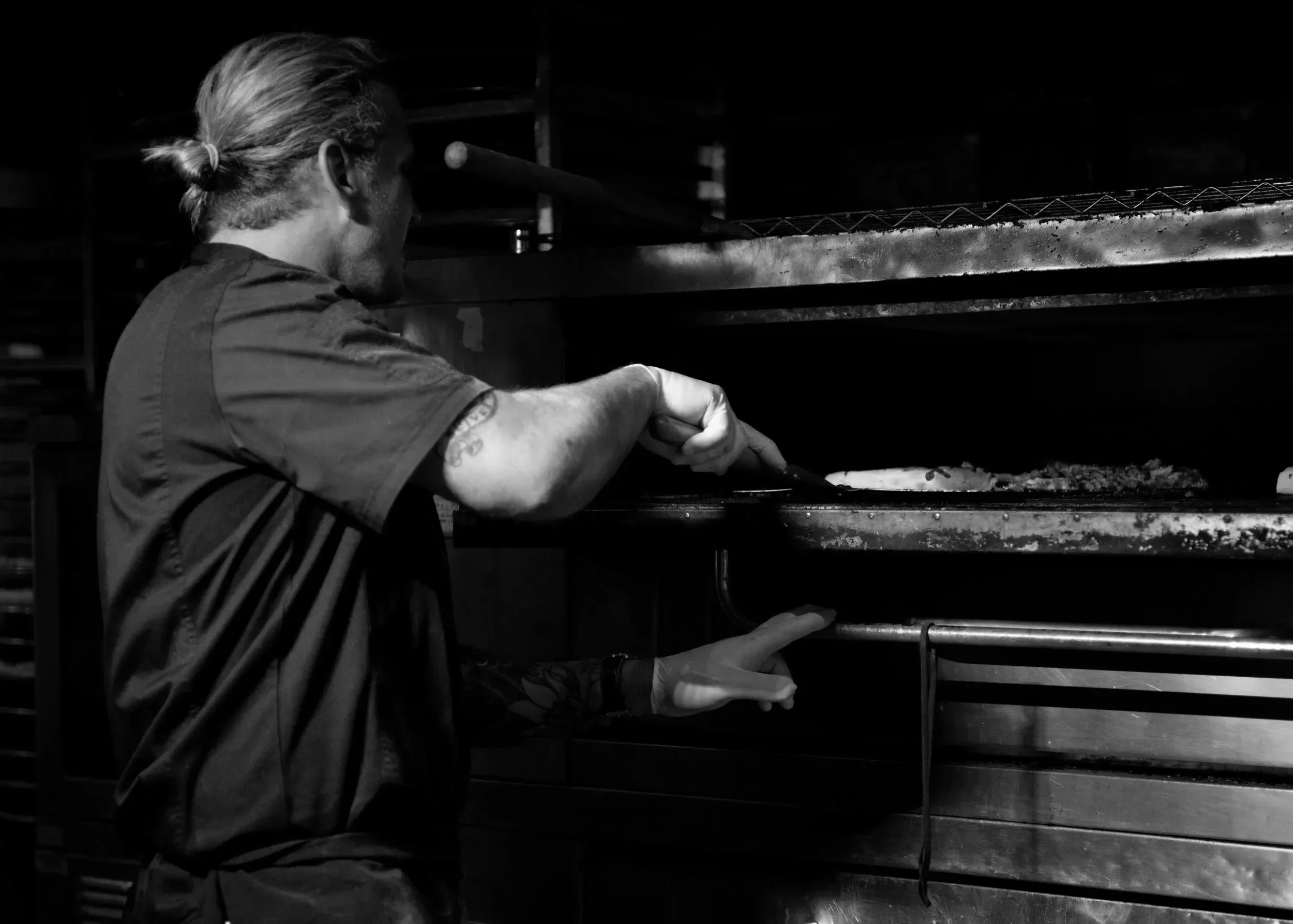 A man with long hair tied back, wearing a short-sleeved shirt, is using a pizza peel to insert or remove a pizza from a commercial pizza oven in a kitchen.