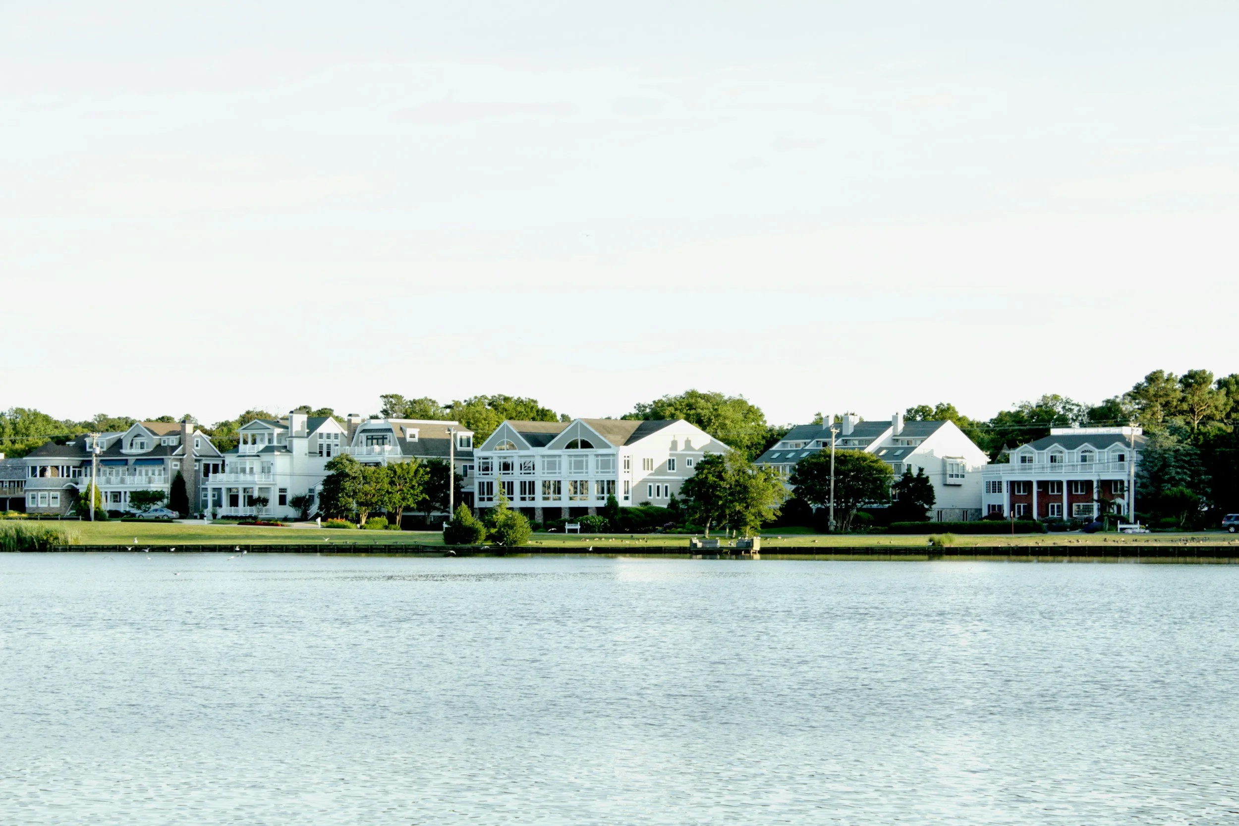 A row of large, white and blue waterfront houses with green trees in front, along a calm body of water under a light sky.