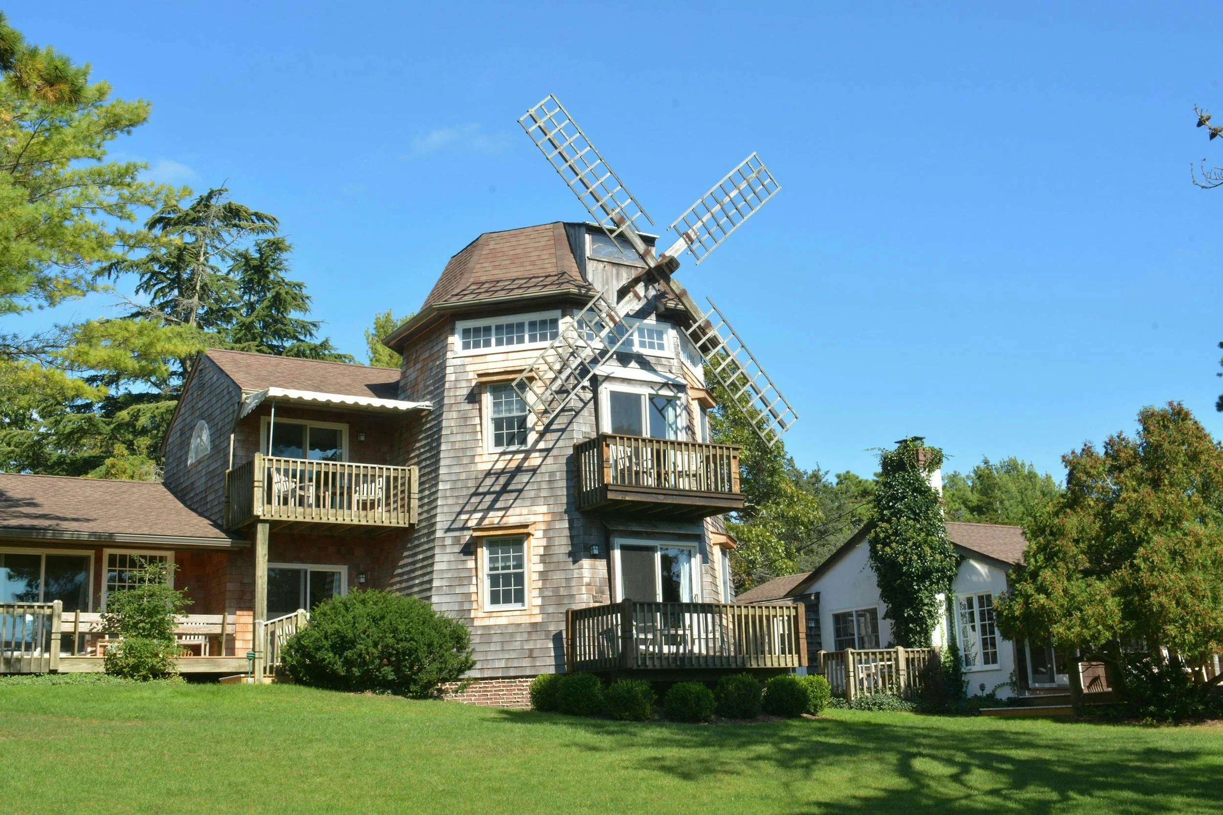 A house with a traditional windmill on top, surrounded by trees and a well-maintained lawn under a clear blue sky.