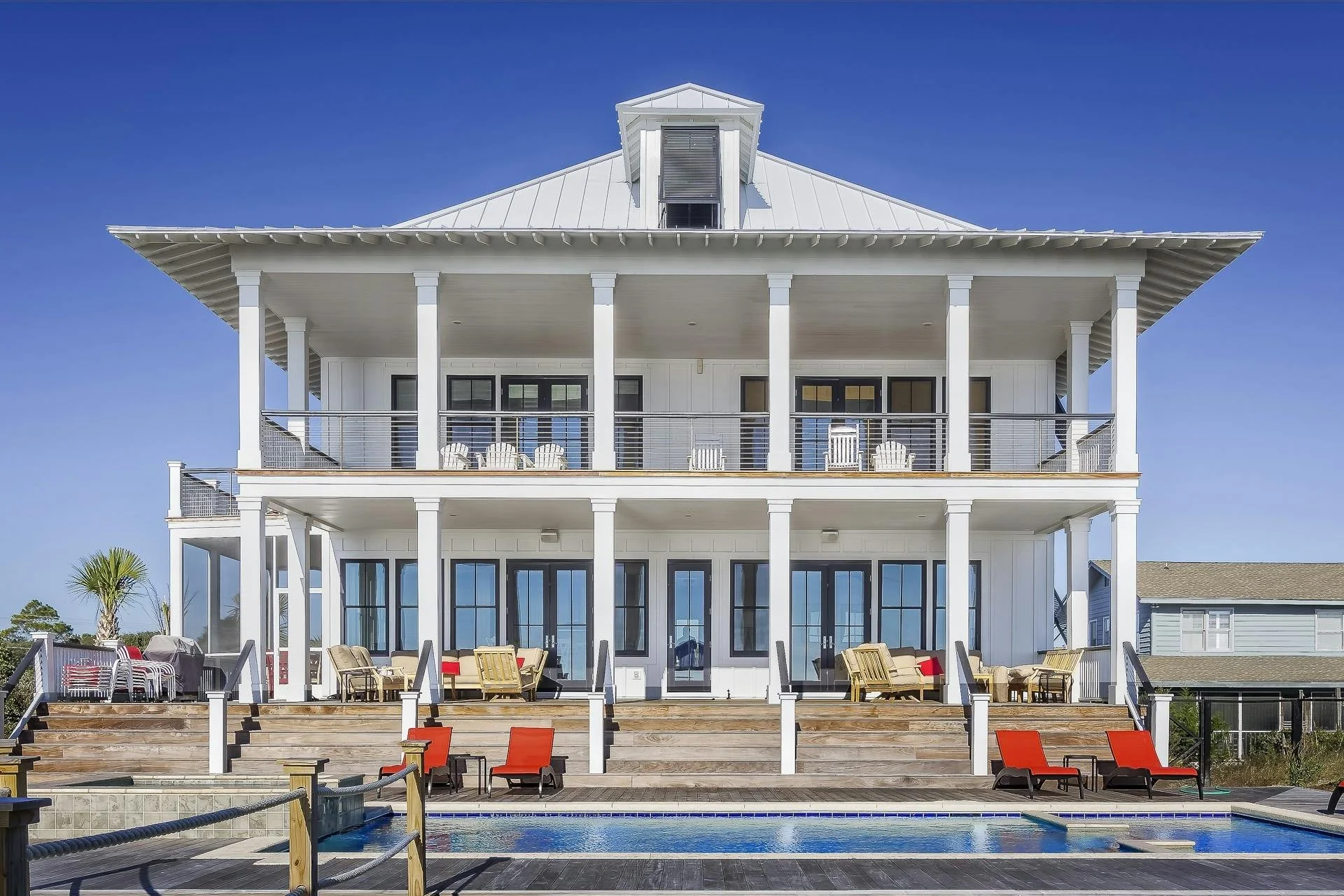 A large, multi-story white house with a metal roof, balconies on each floor, and glass doors and windows. There are outdoor chairs and lounge chairs on the deck, with a swimming pool in the foreground. Clear blue sky overhead.
