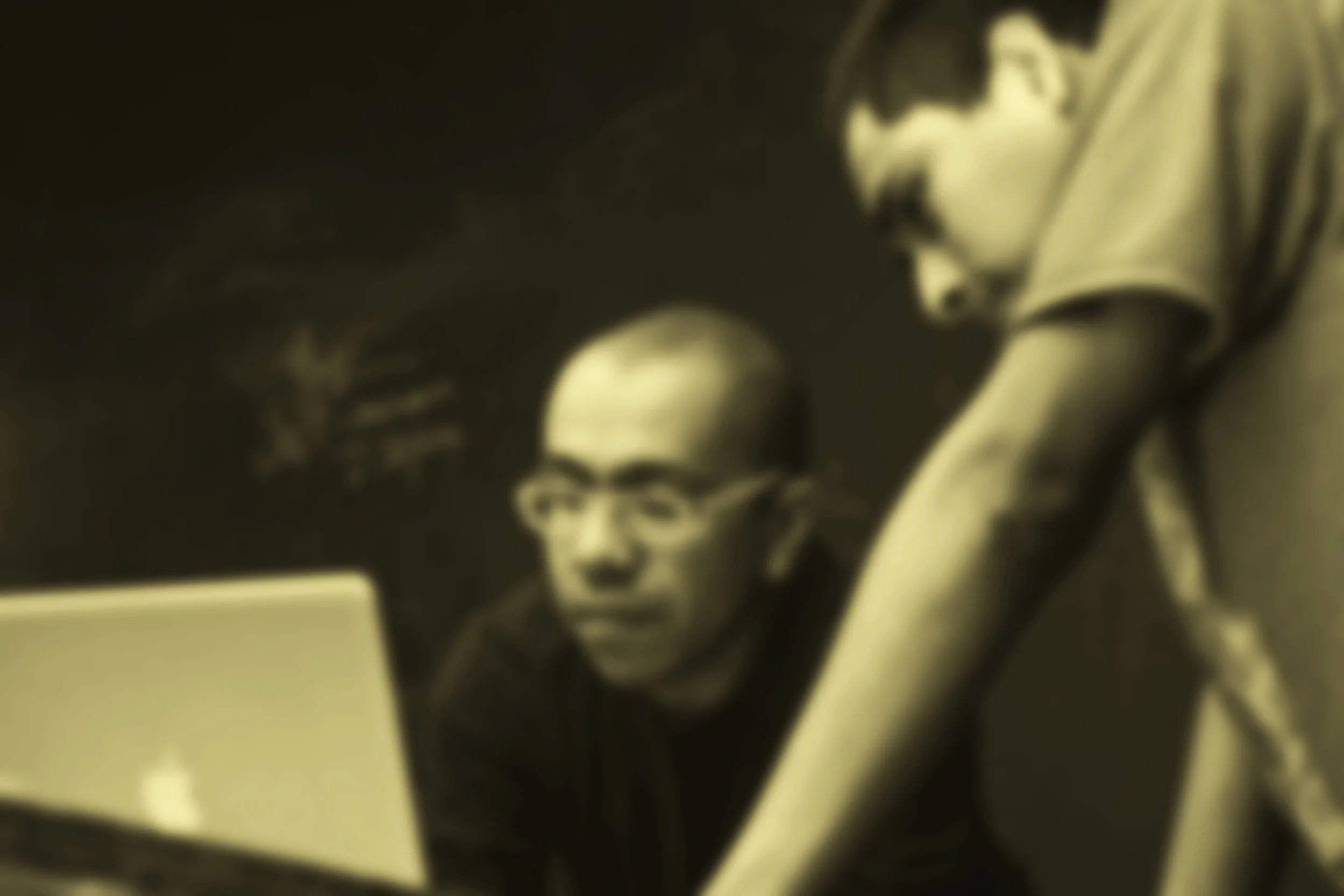 A teacher with glasses working on a laptop, with a student leaning over to look at something on the screen, in a classroom setting.