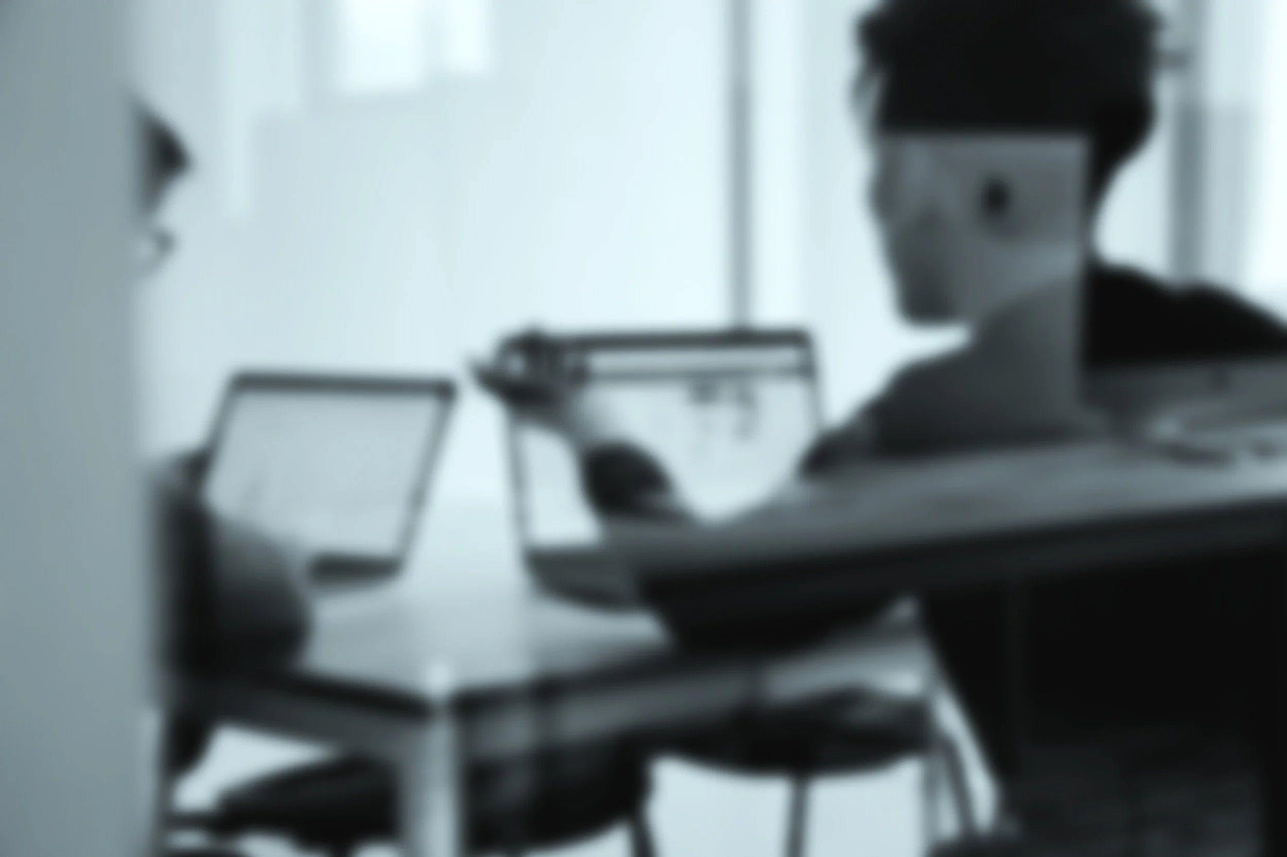 A person with short hair and glasses working at a desk in front of a laptop, in a blurry black and white photo.