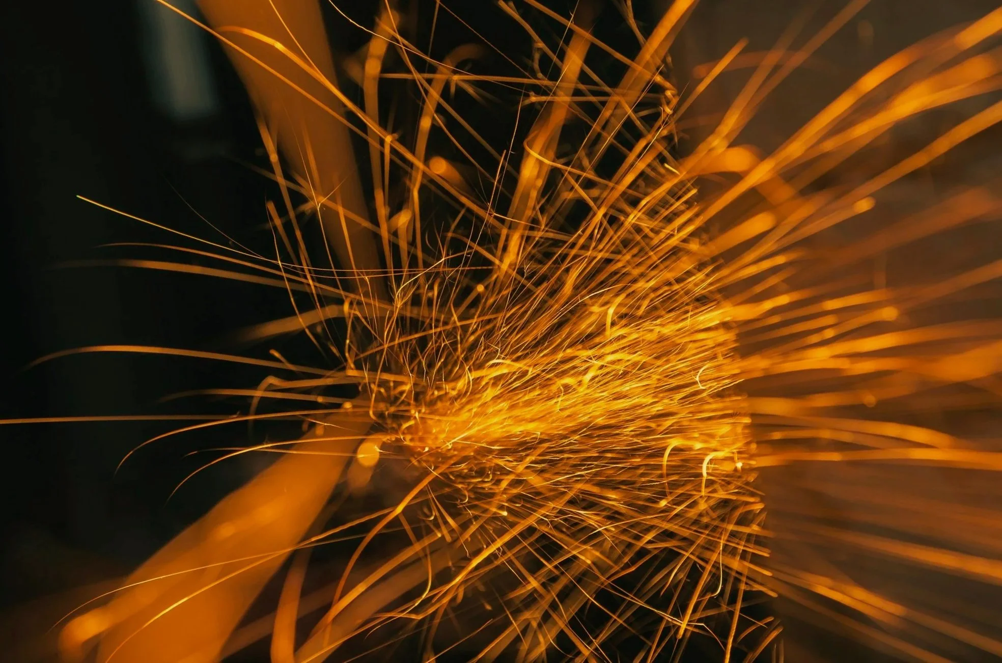 Close-up of sparks flying from a firework or metal work, with bright orange lines radiating outward against a dark background.