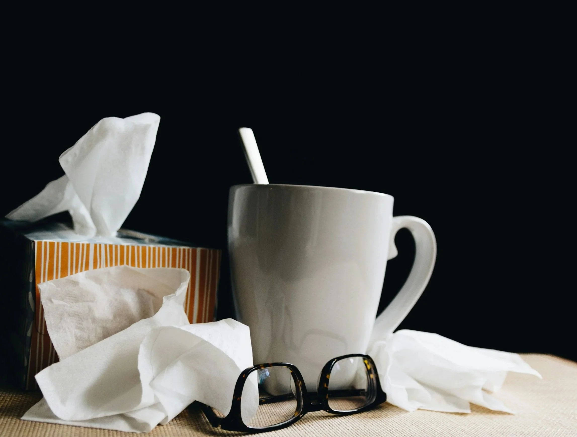 A white ceramic mug with a spoon inside, surrounded by crumpled tissues, a box of tissues, and a pair of black glasses, on a surface against a dark background.