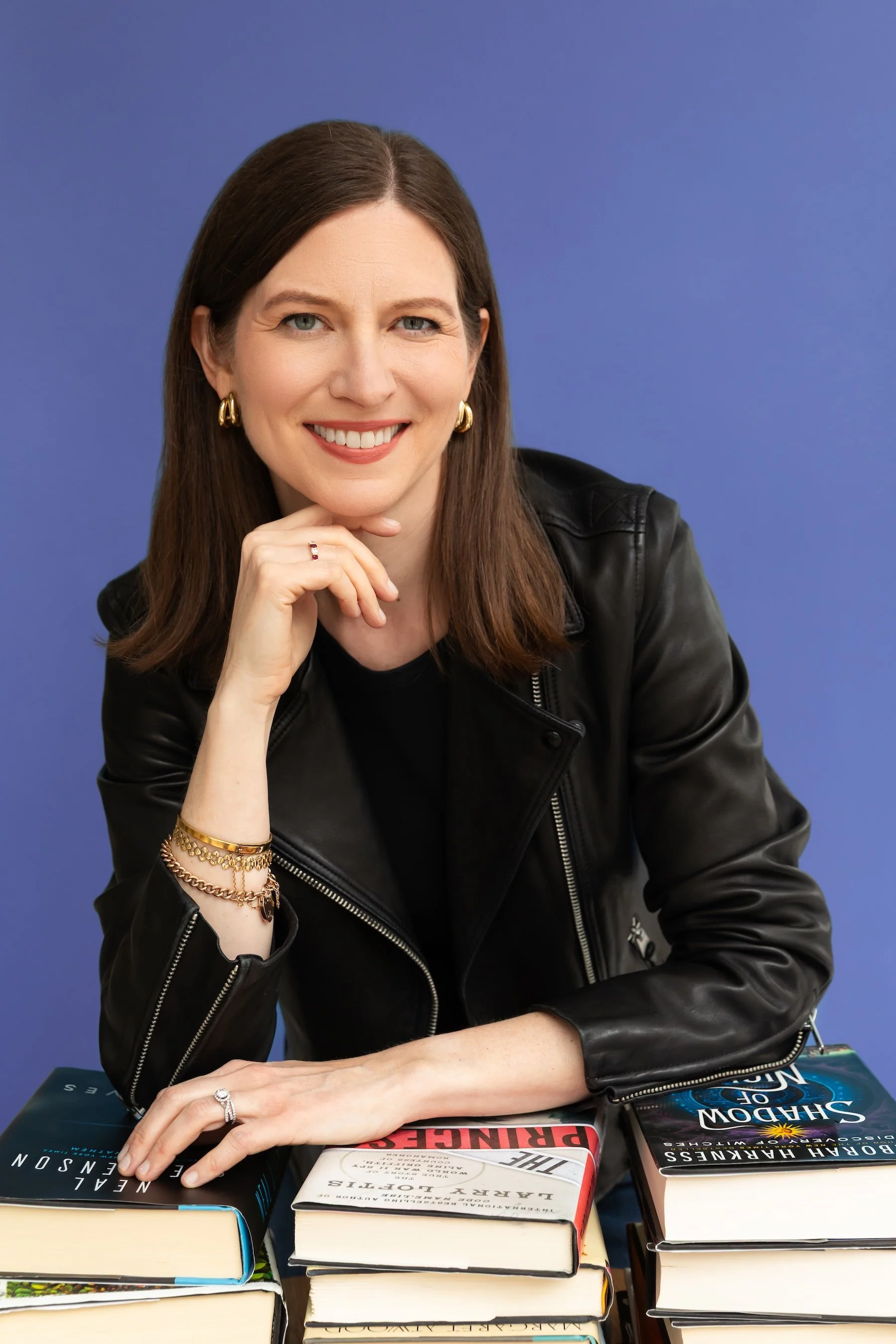 Woman with brown hair, wearing gold jewelry and a black leather jacket, sitting at a table with multiple books, smiling and resting her chin on her hand against a blue background.