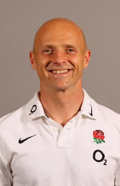 Portrait of a smiling man with a shaved head wearing a white sports polo shirt with English rugby team logos and sponsors.