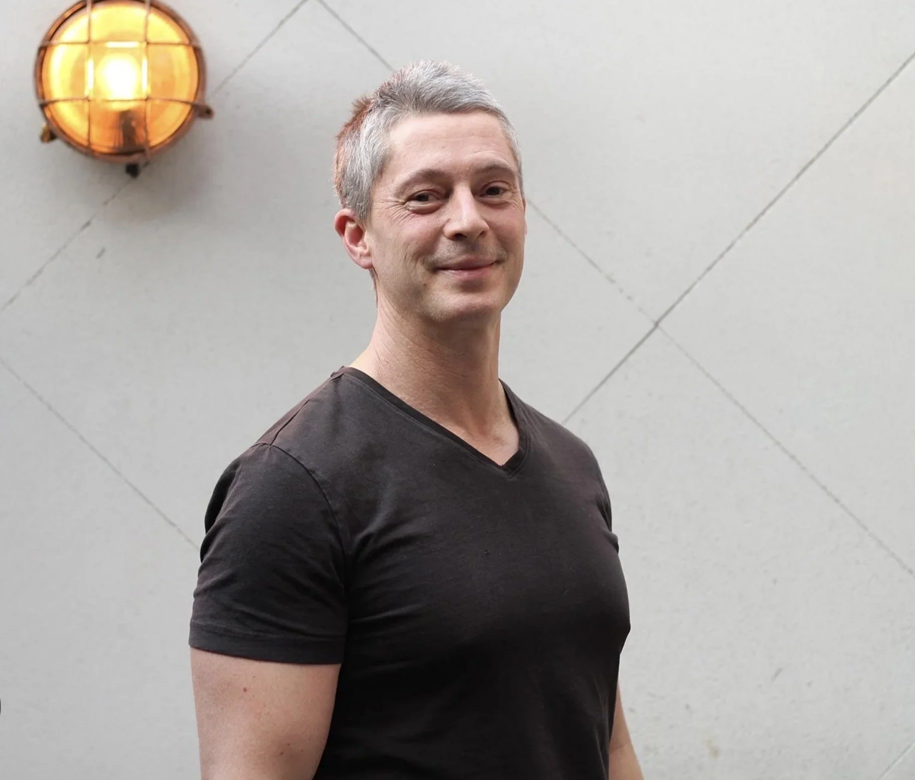 A man with short gray hair wearing a black t-shirt, standing outdoors in front of a gray wall.