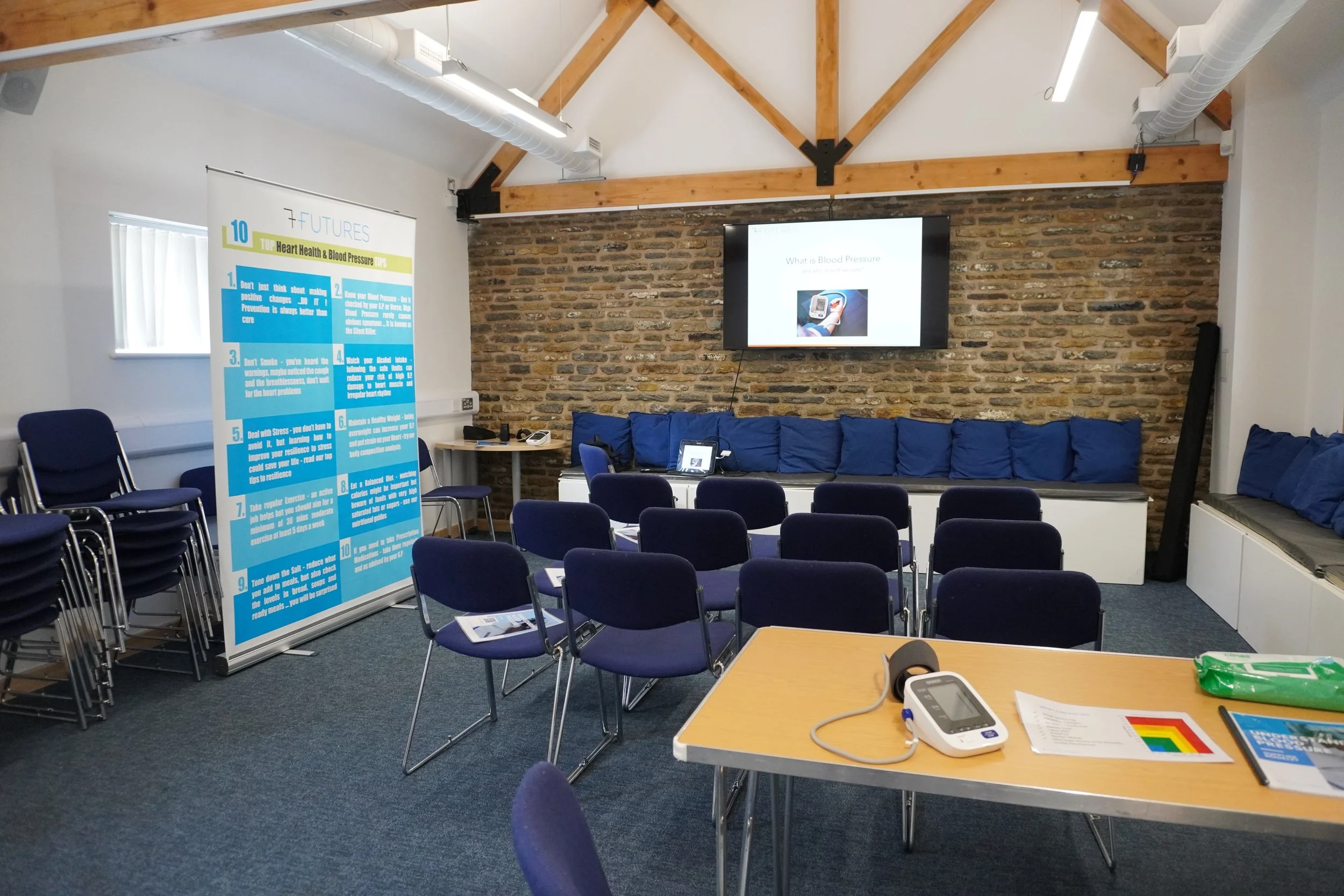 A meeting or presentation room with chairs facing a wall-mounted TV screen displaying a slide titled 'What is Blood Pressure?'. There are stacked chairs to the left, a table with a blood pressure monitor and some papers in the foreground, and a large blue and white educational banner on the left side of the room. The room has a brick wall at the back, a wooden beam ceiling, and a small window with vertical blinds.