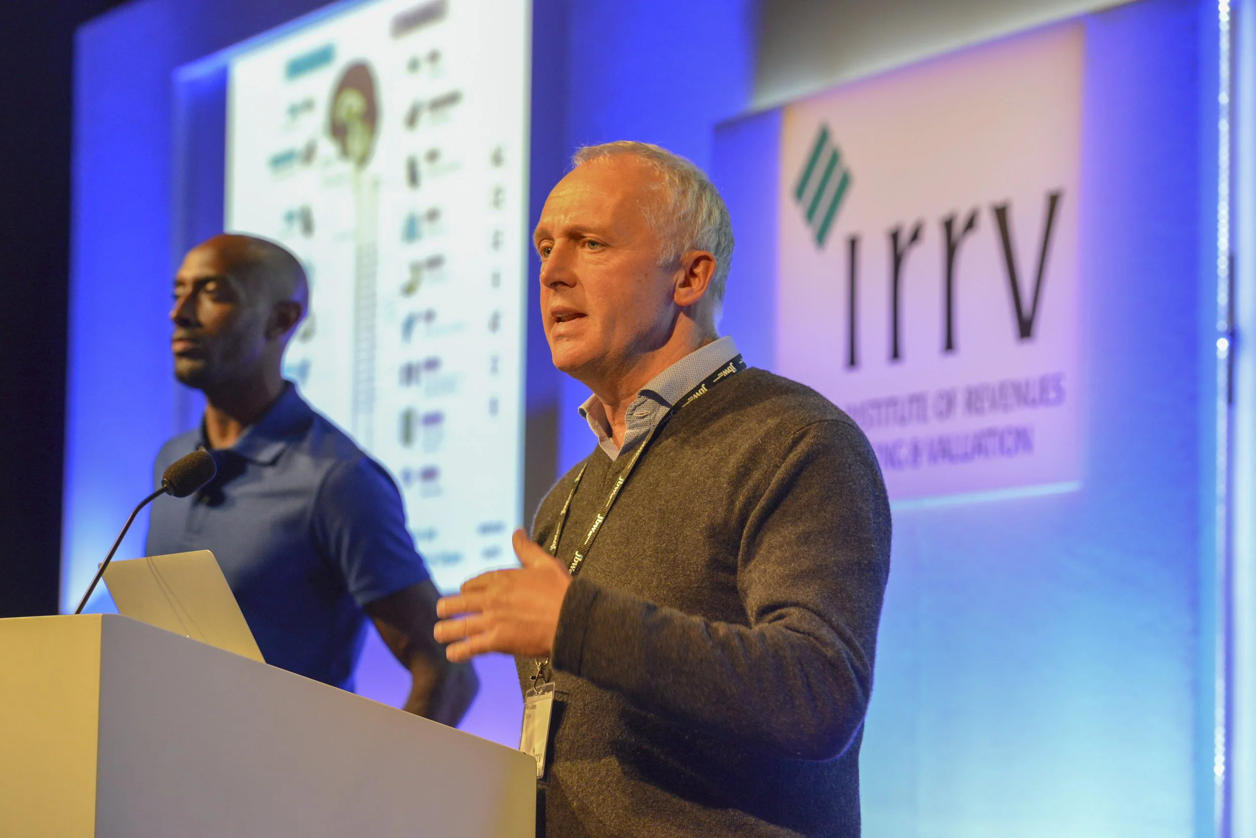 Two men speaking at a conference, with a presentation screen in the background displaying a chart and the IRV logo.