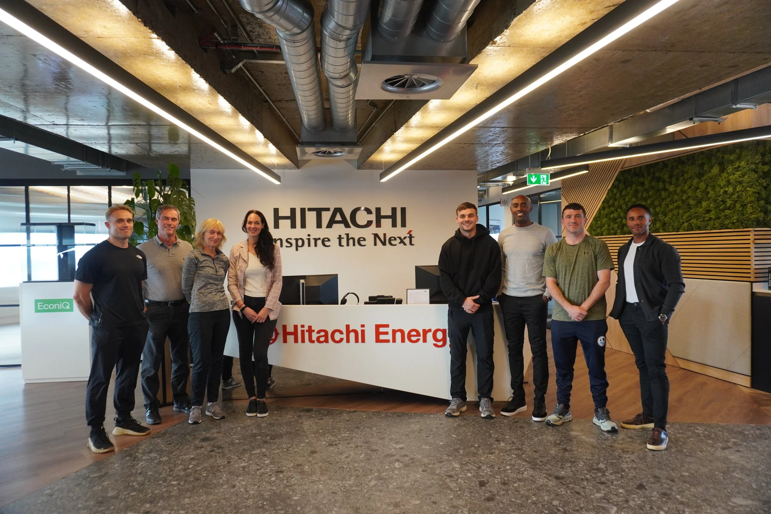 Group of nine people standing in front of a HITACHI sign and reception desk inside an office building.