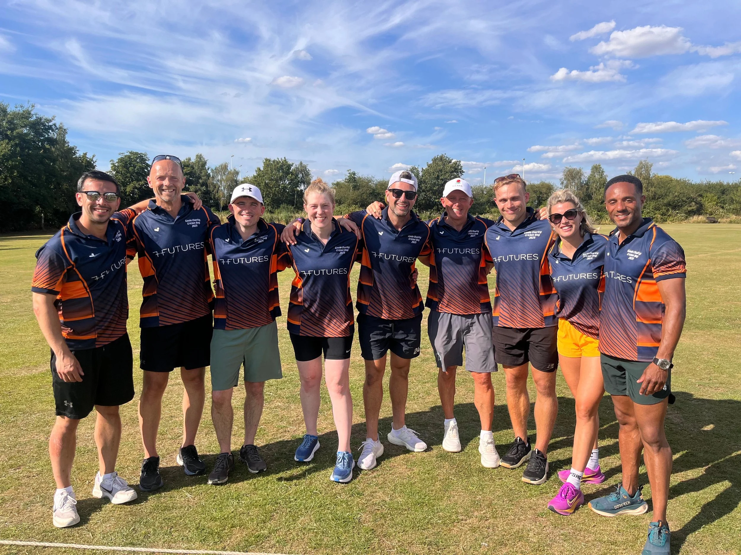 Group of nine people standing outdoors on grass field, smiling, with trees and blue sky in the background, wearing matching sports jerseys.