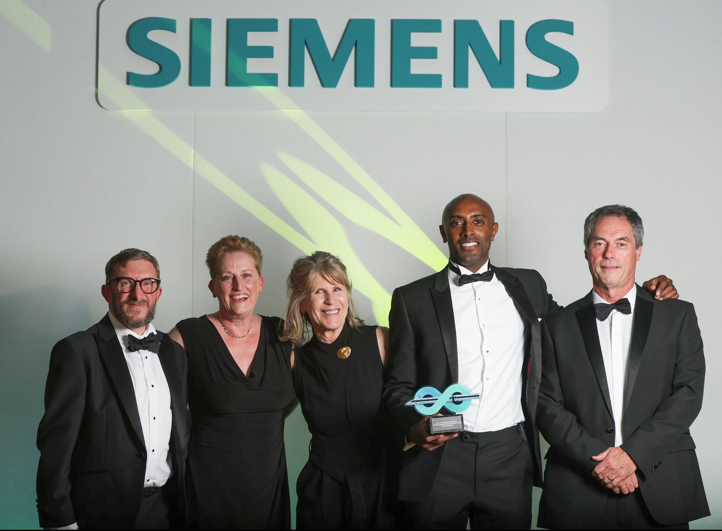 Group of five people in formal attire standing in front of a wall with Siemens logo, with one person holding an award. They are smiling and posing for the photo.